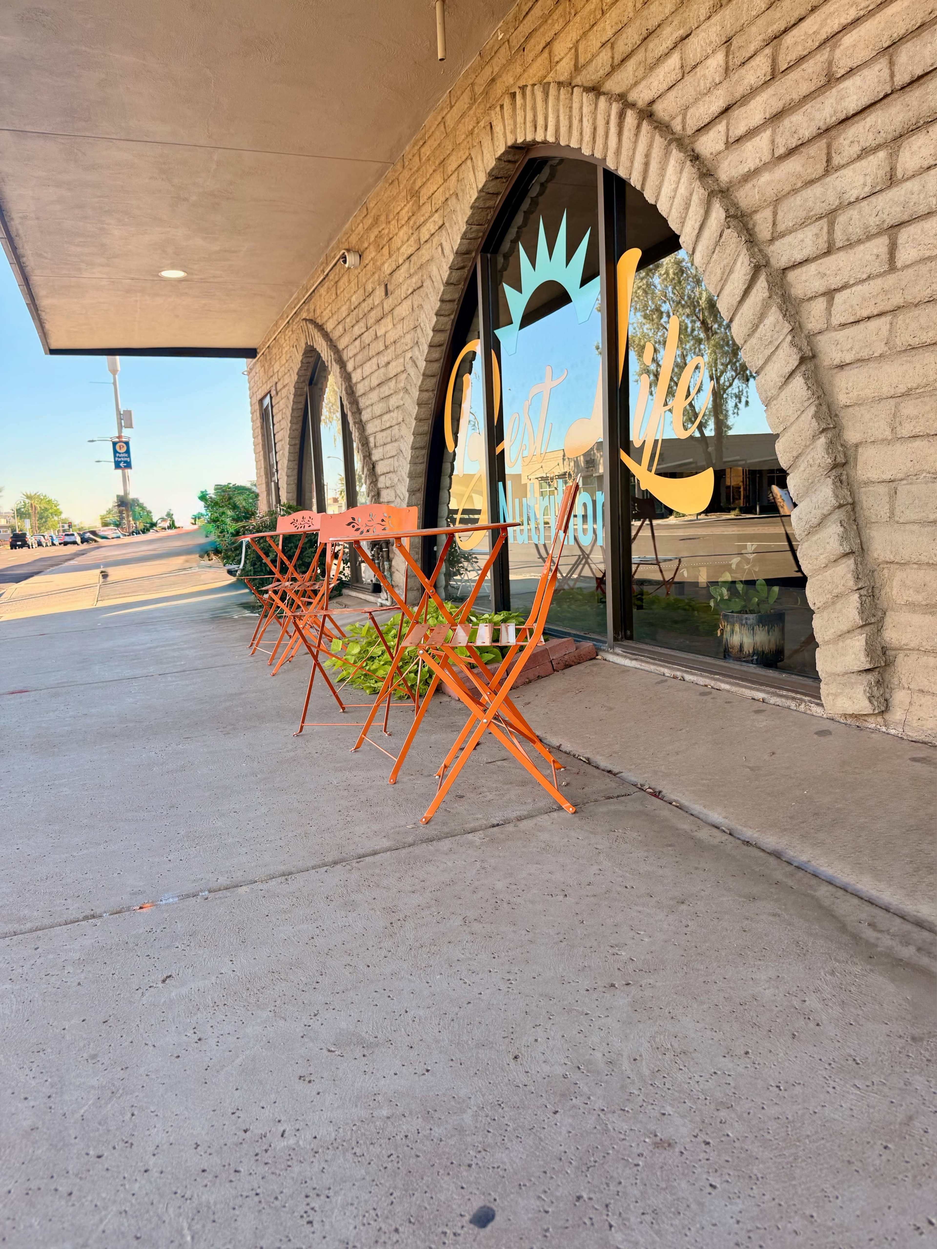 The image shows a sidewalk in front of a storefront with arched windows and a couple of bright orange chairs lined up outside.