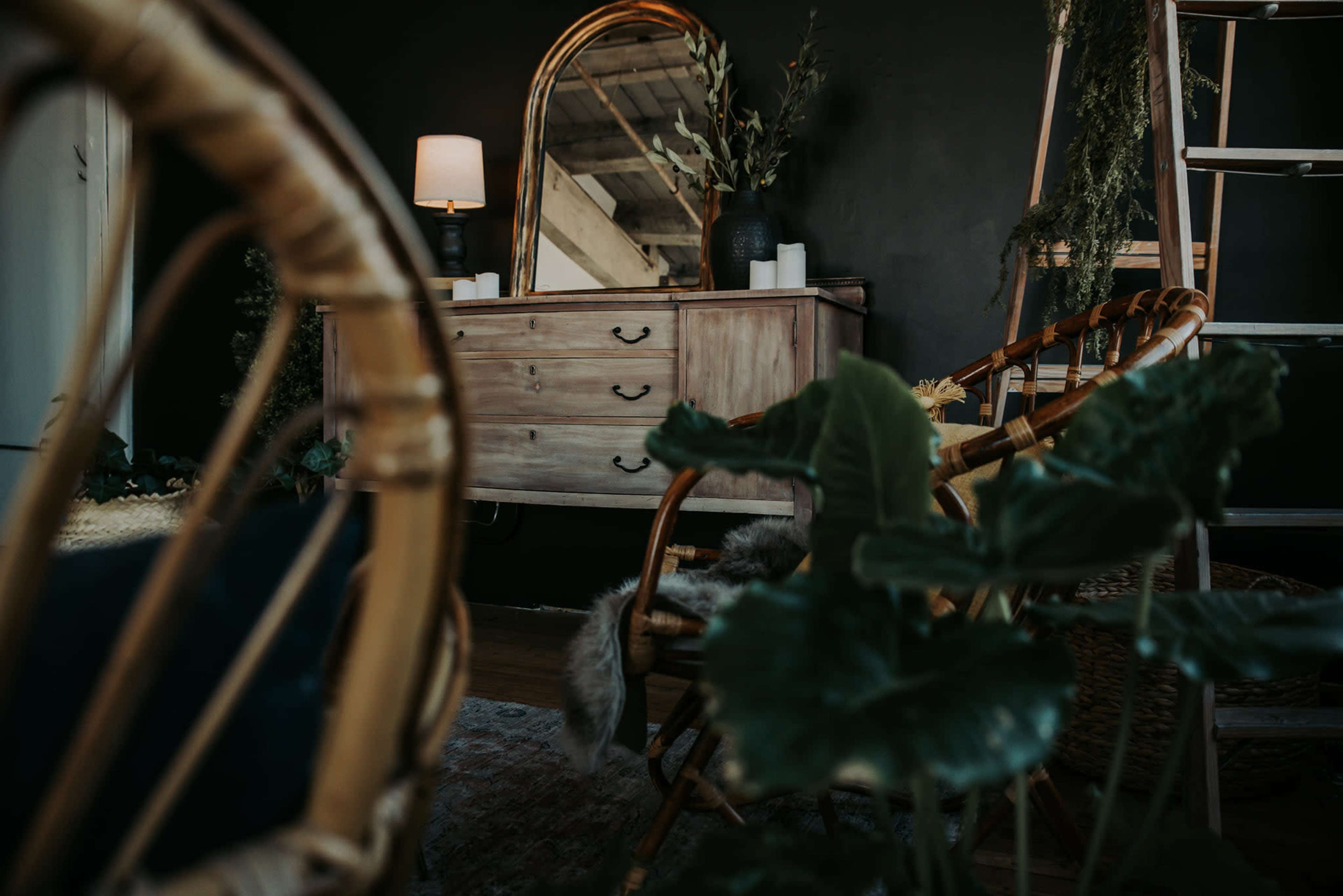 A cozy room featuring a wooden dresser, a round mirror, a lamp, and wicker furniture surrounded by greenery.