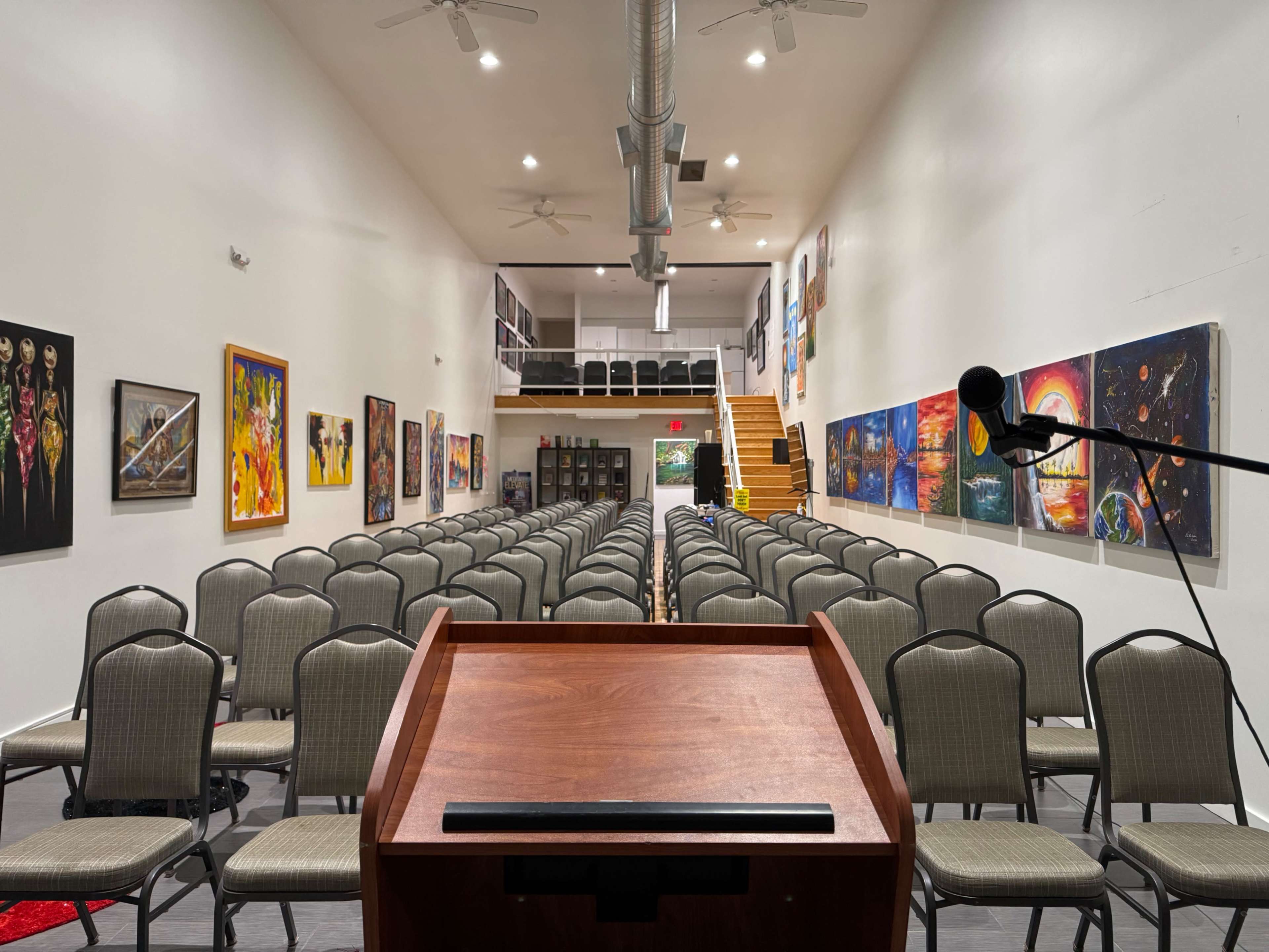 A dimly lit auditorium features rows of gray chairs facing a wooden podium, with colorful artwork displayed on the white walls.