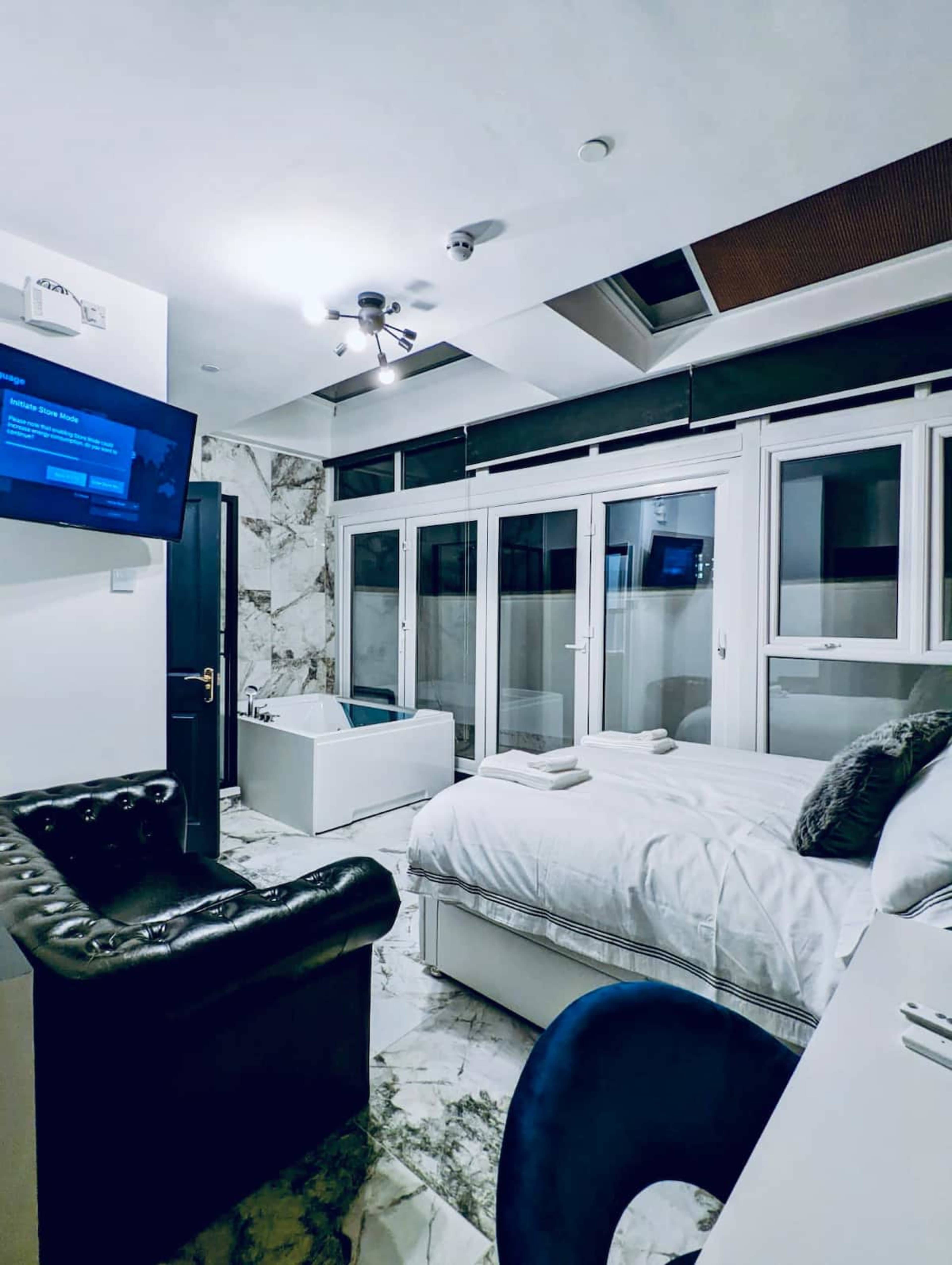 A modern bedroom with a bed, a black leather chair, a bathtub, and large windows letting in natural light.