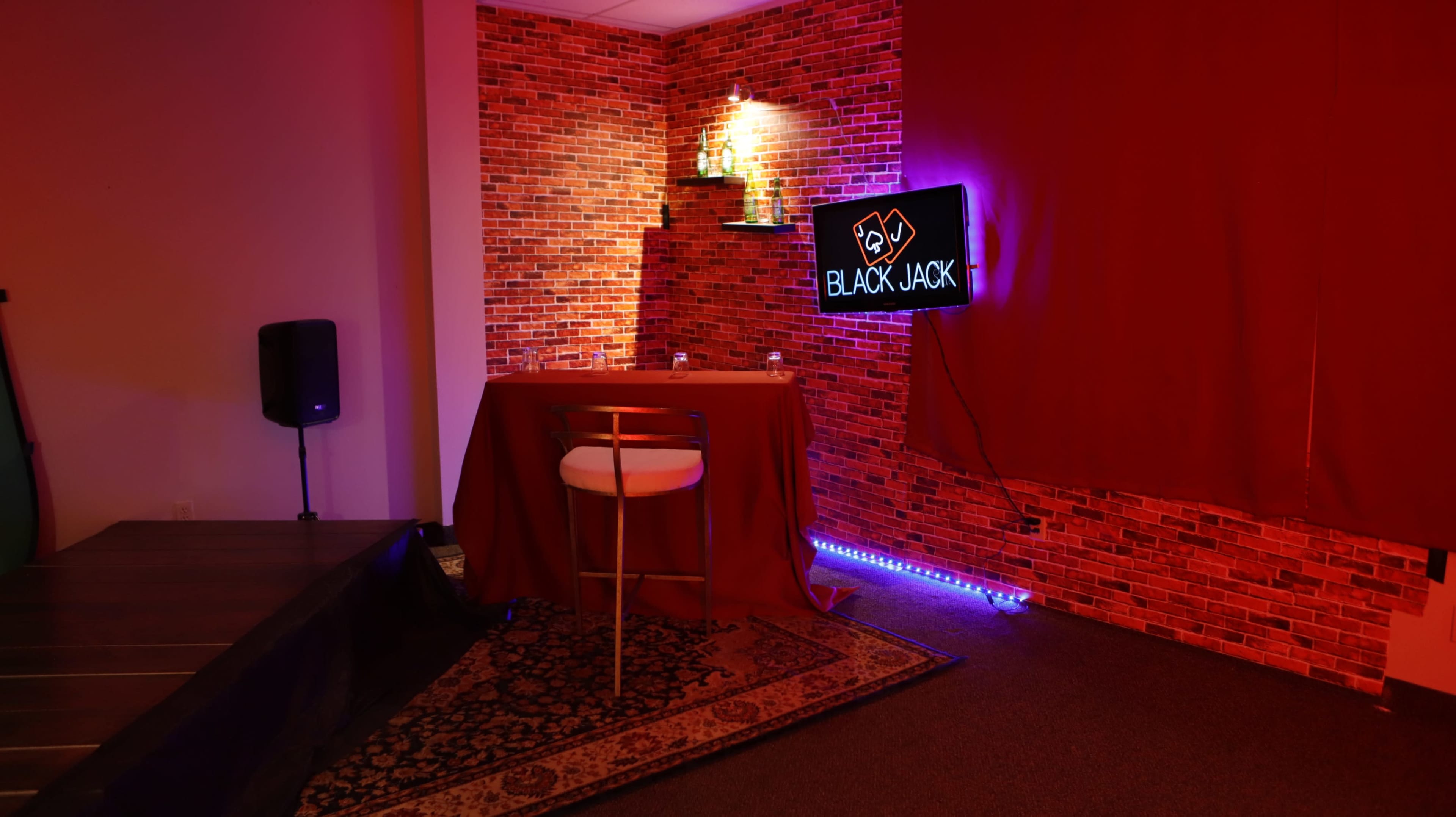 A small room features a red-brick wall, a neon "BLACKJACK" sign, and a single table with a white chair, illuminated by ambient lighting.
