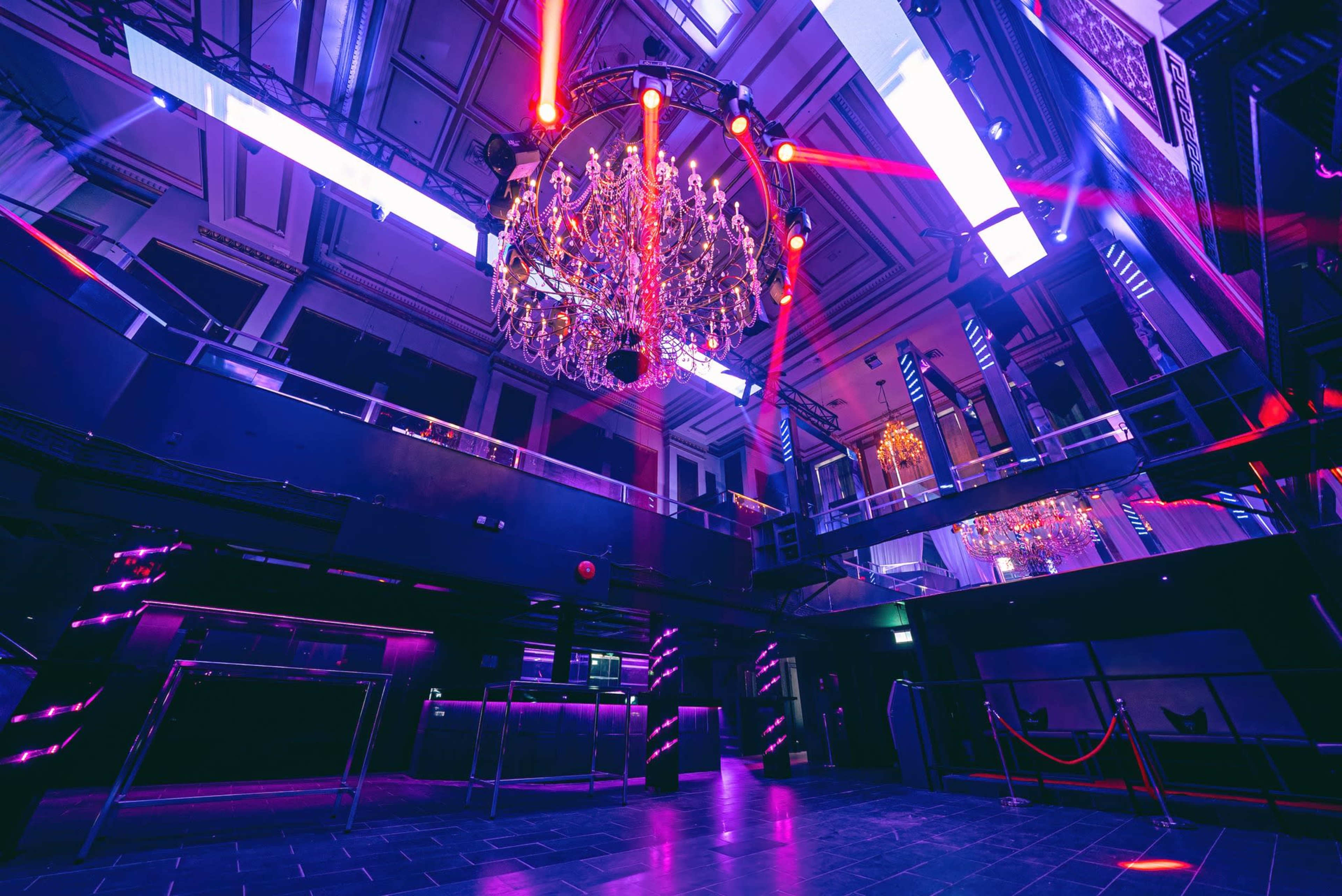 The image shows a spacious nightclub interior with a large chandelier and vibrant lighting effects, featuring multiple levels and a dark floor.