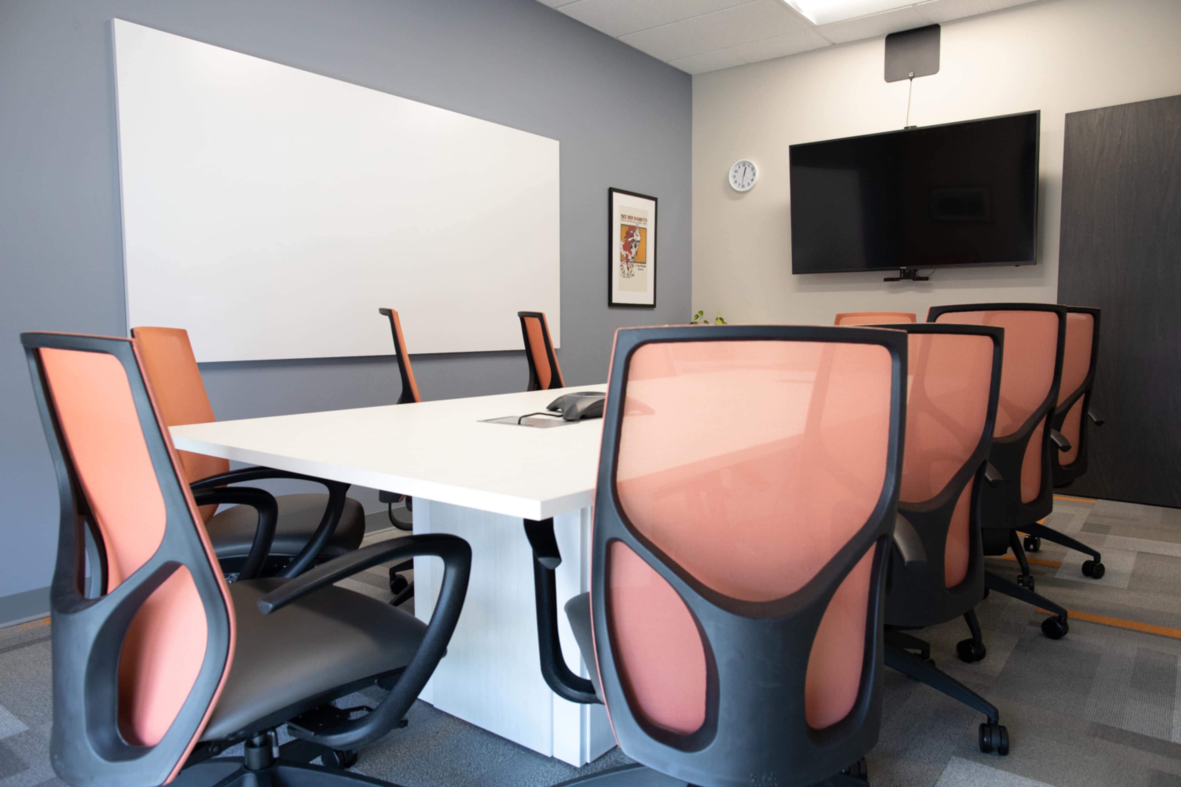A conference room features a large white table surrounded by eight office chairs with orange backs, a wall-mounted television, and a whiteboard.