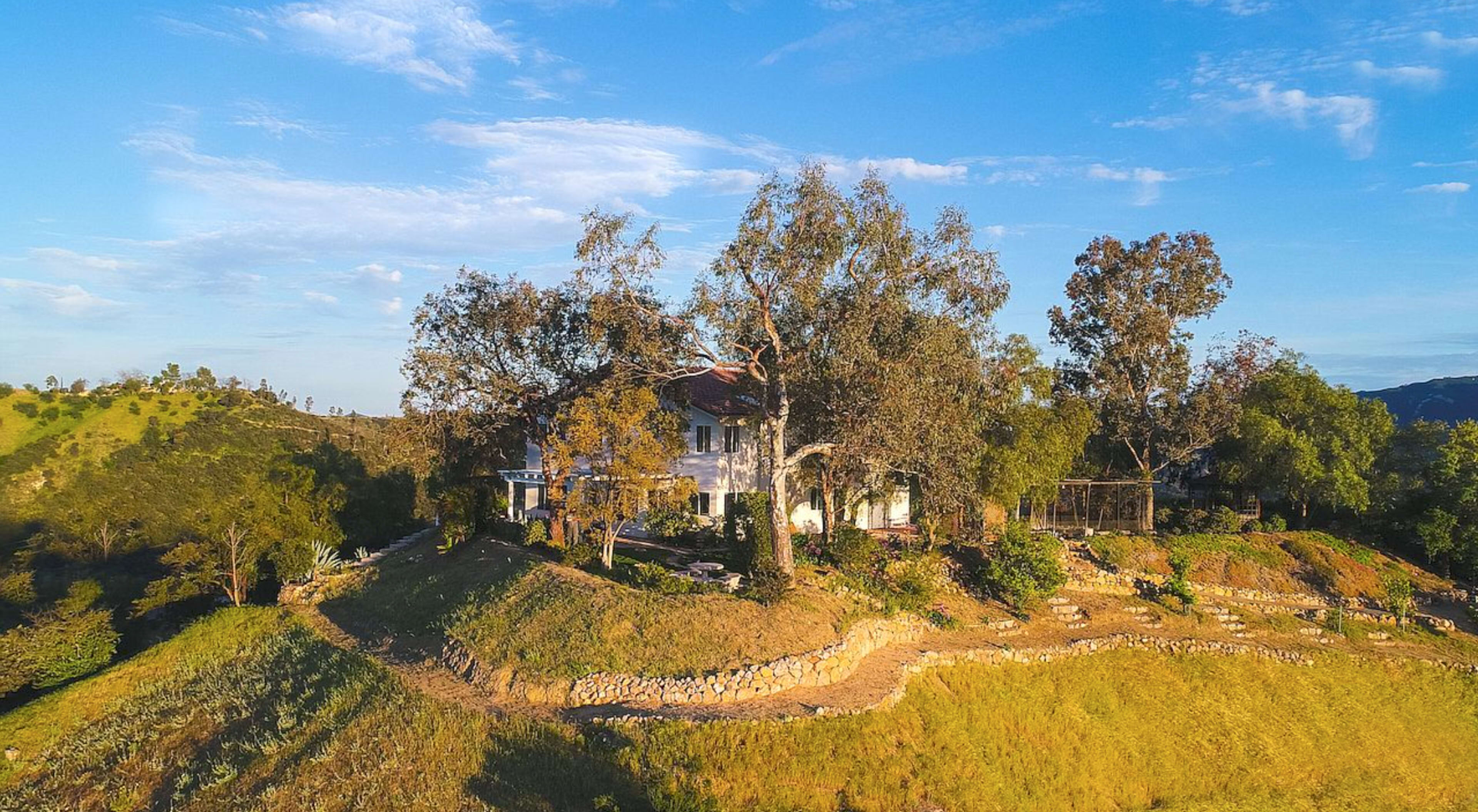 A white house is situated on a hillside, surrounded by trees and a winding path leading to it.