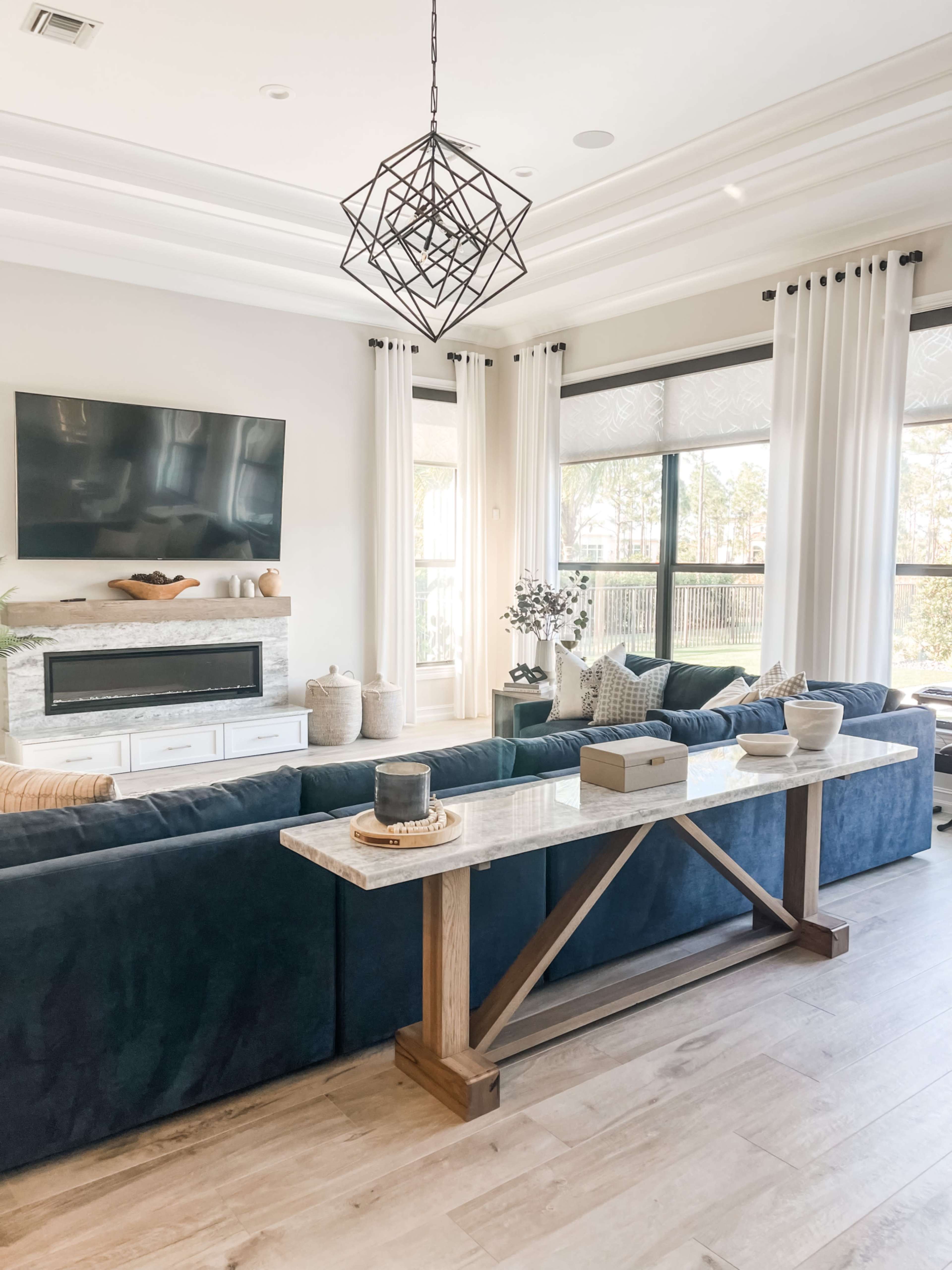 A modern living room features a blue sofa, a marble-top console table, a mounted television, and large windows covered with light curtains.