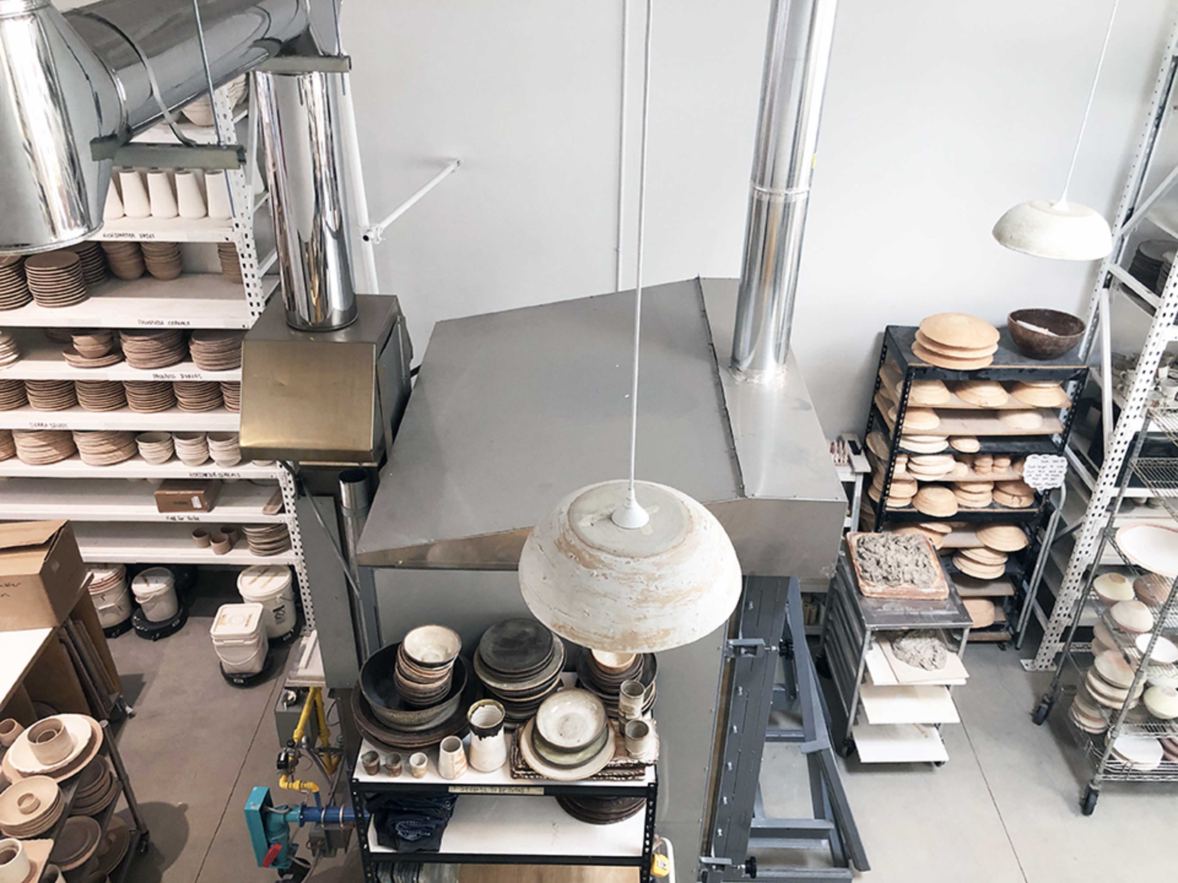 A workshop space with shelves of ceramic plates and bowls, metal ducts, and overhead lighting.