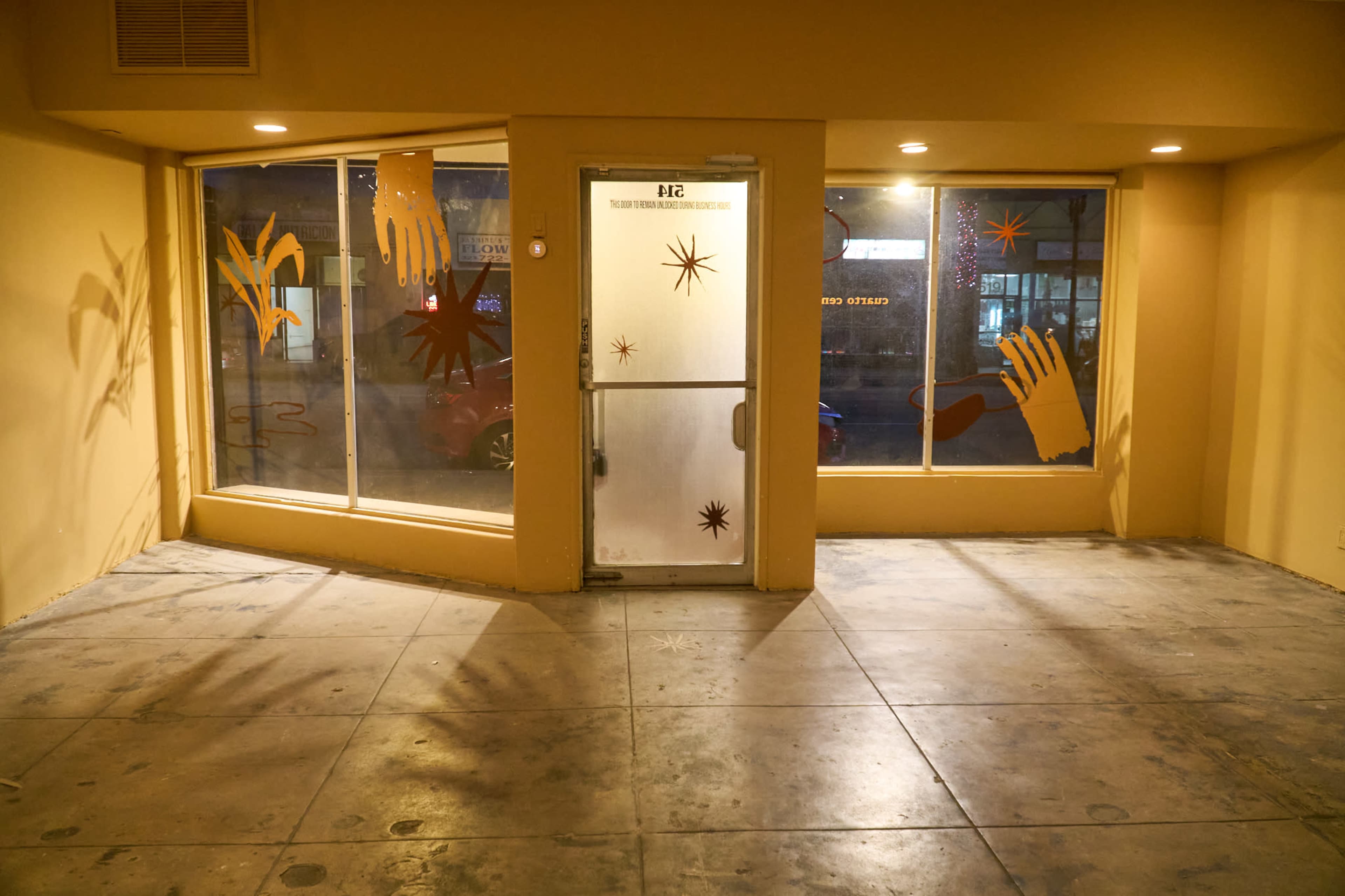 The image shows an empty room with yellow walls, a frosted glass door, and large windows featuring colorful hand decorations.