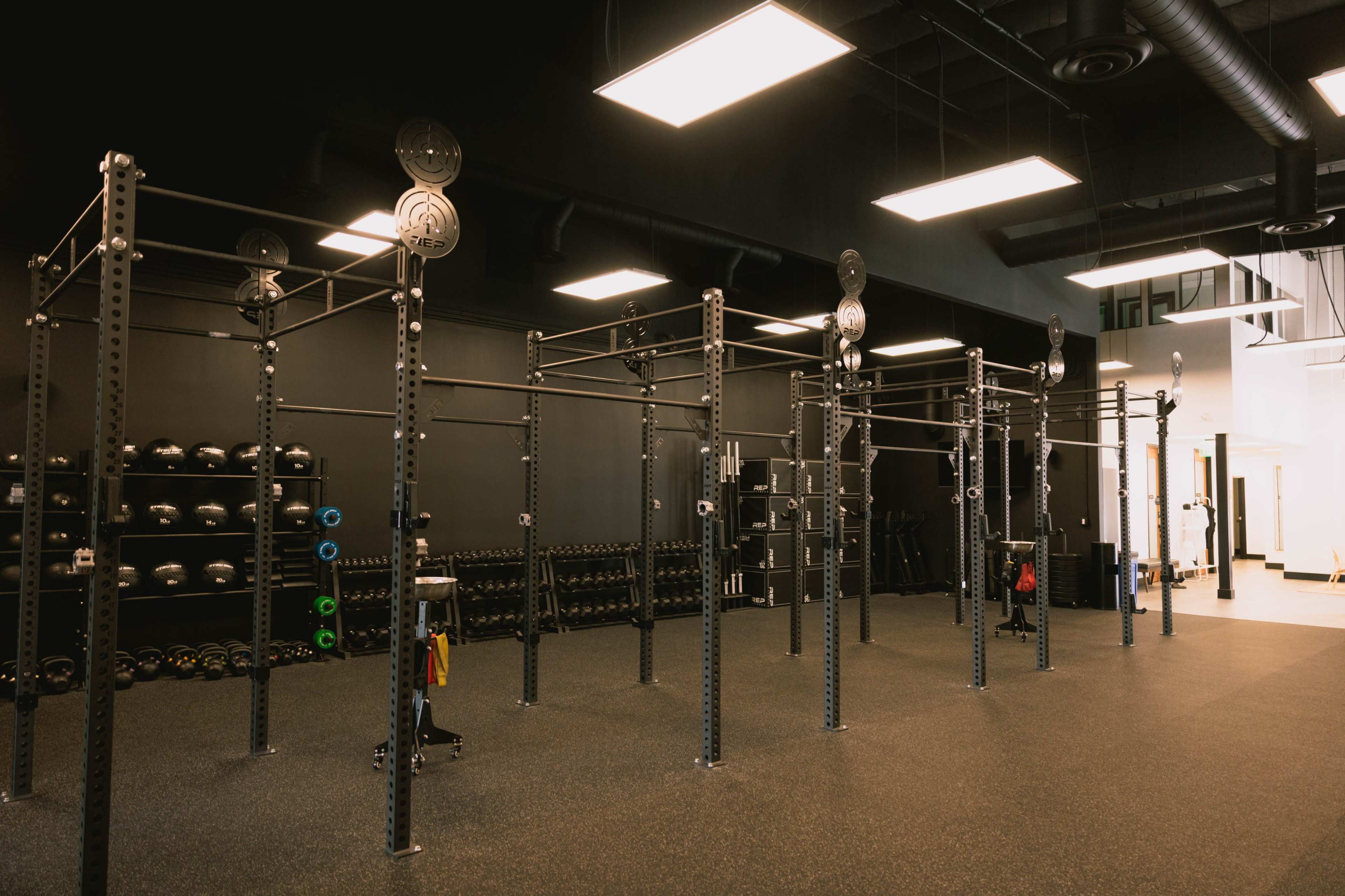 The image shows a spacious gym interior with various weightlifting equipment, including racks for barbells and a section for free weights on shelves against the wall.