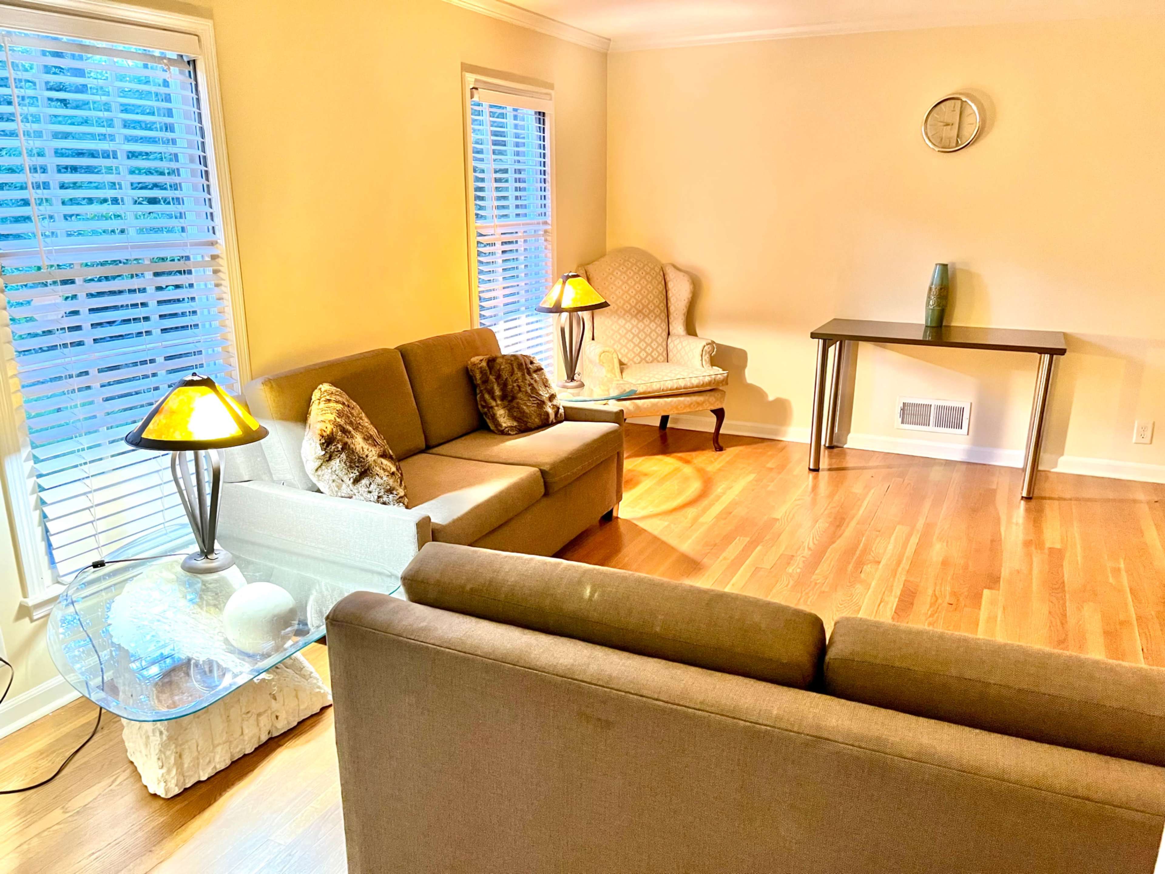 The image depicts a living room with two sofas, a glass coffee table, a side chair, and a table situated against a wall with windows and a clock.