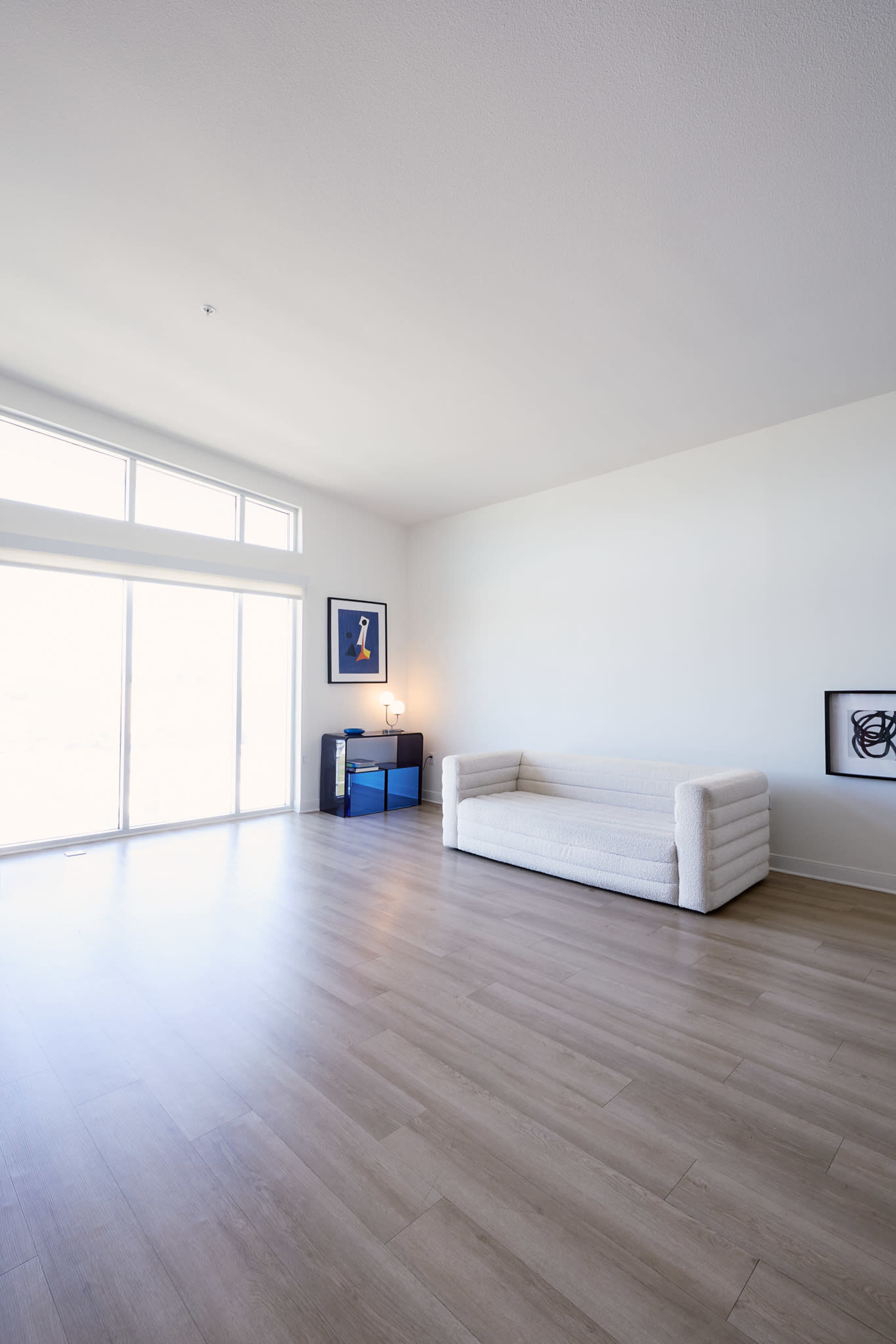 A spacious, minimalistic living room features a white couch, a blue side table, and large windows allowing natural light to enter.