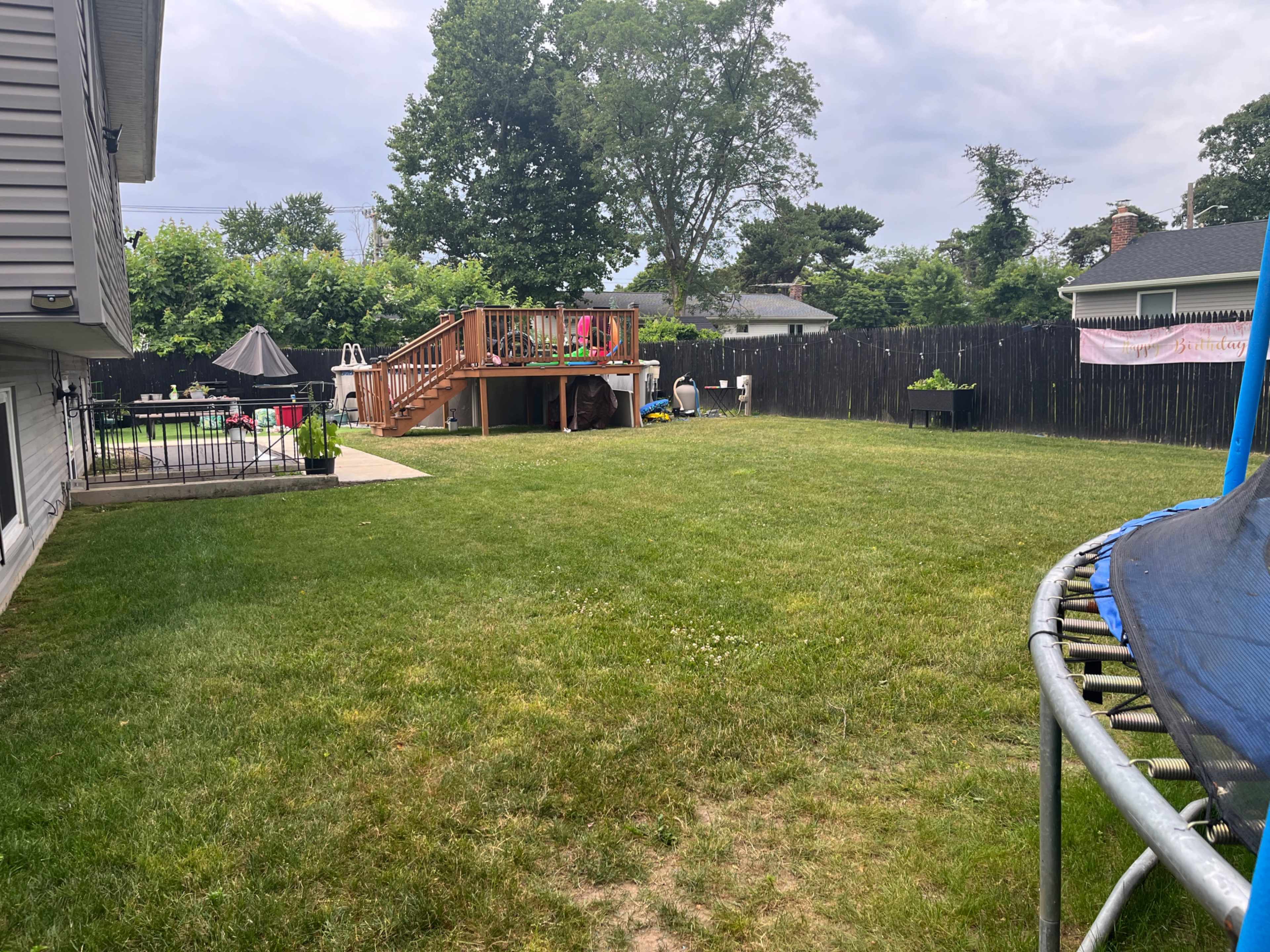 The image shows a backyard with a grassy area featuring a trampoline, a wooden deck, outdoor furniture, and a black fence in the background.