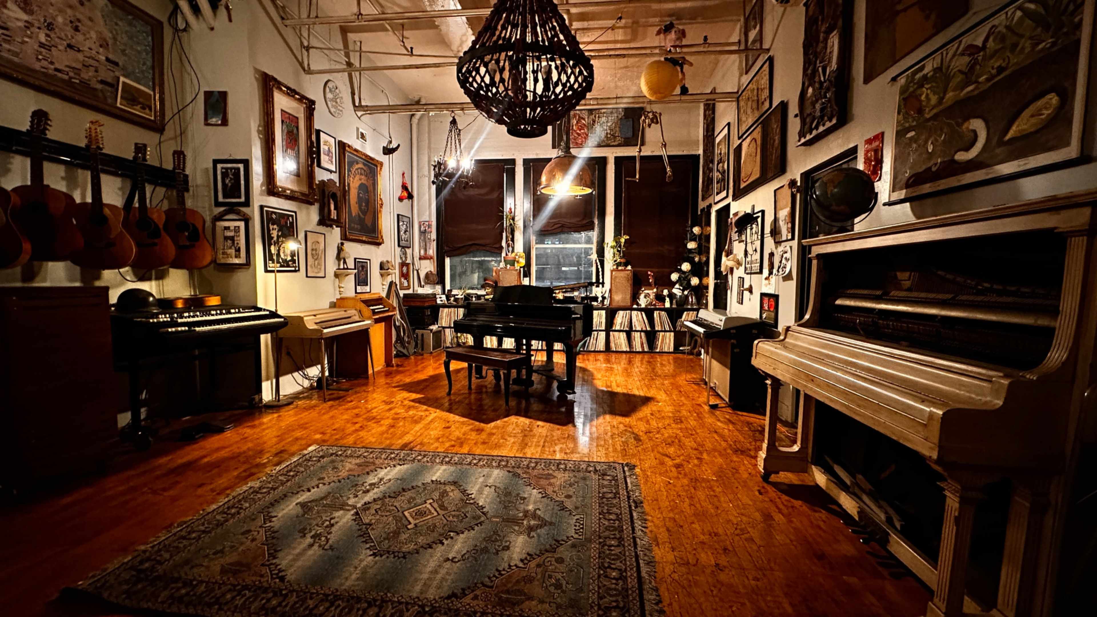 The image shows a warm, artistic music studio featuring a grand piano, vintage keyboards, and various framed artworks on the walls.