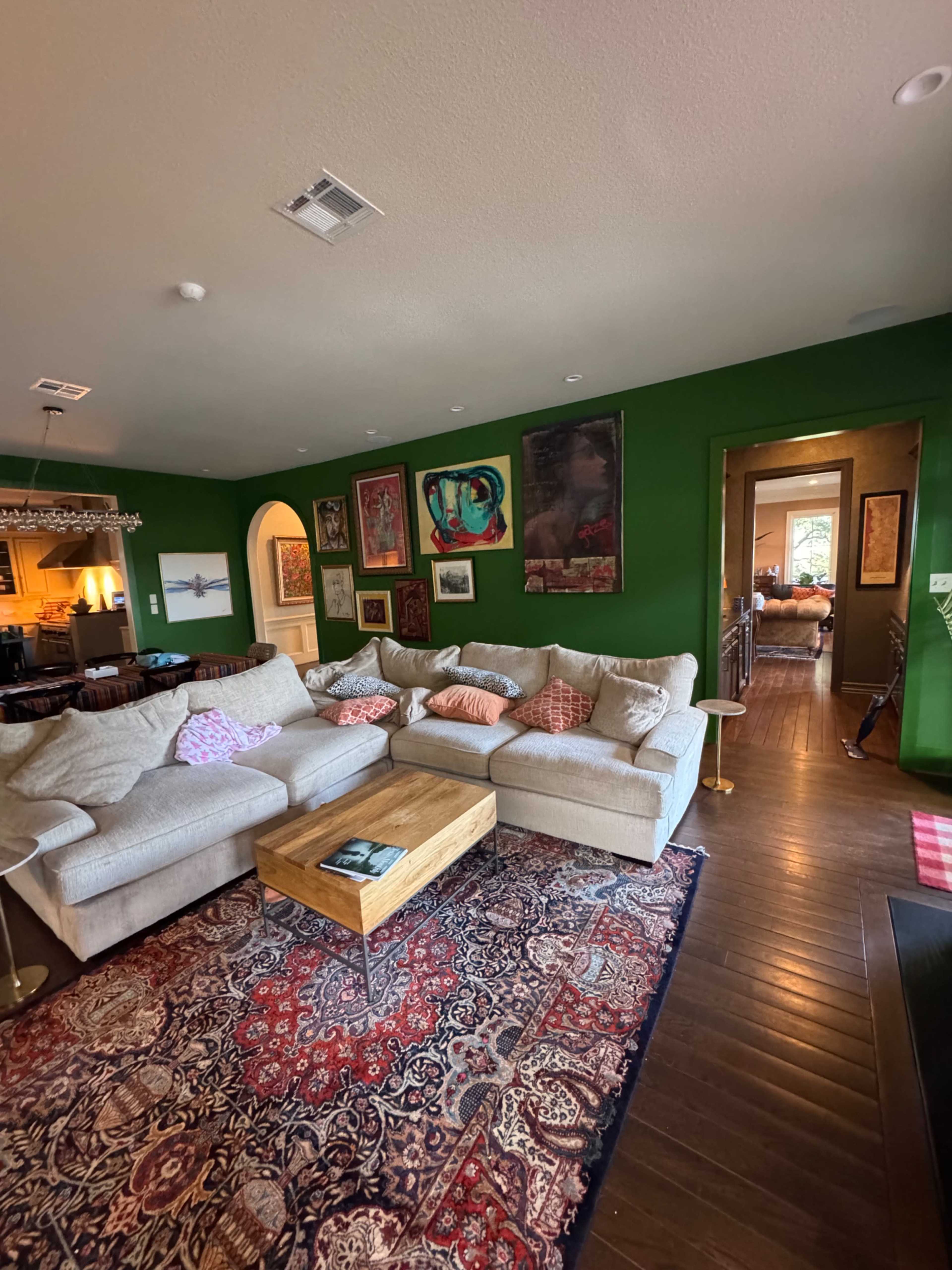 The image shows a living room with a large beige sectional sofa, a wooden coffee table, a colorful area rug, and green walls adorned with various framed artworks.