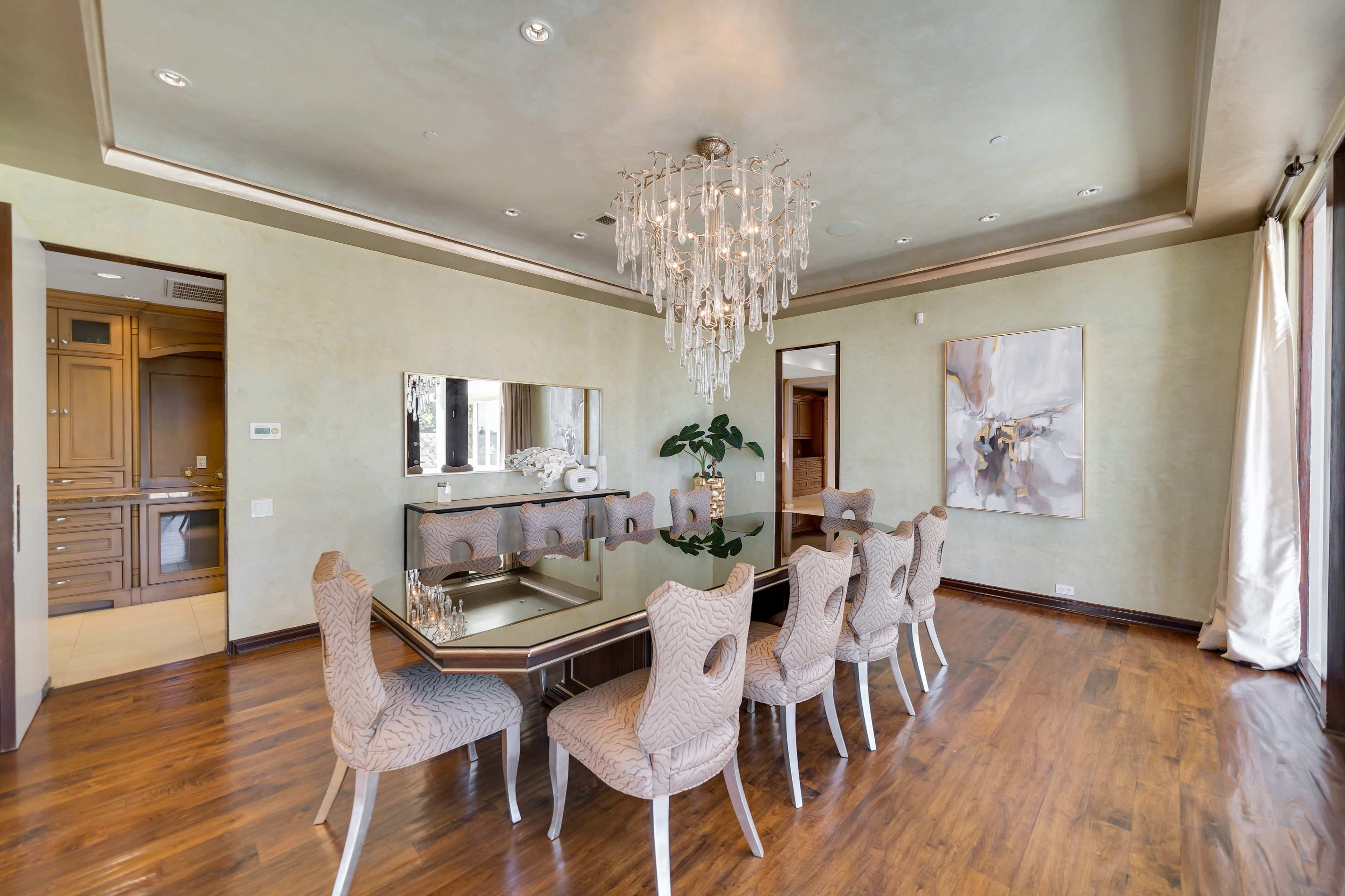 A modern dining room features a large table with eight upholstered chairs, a crystal chandelier, and a decorative mirror on the wall.
