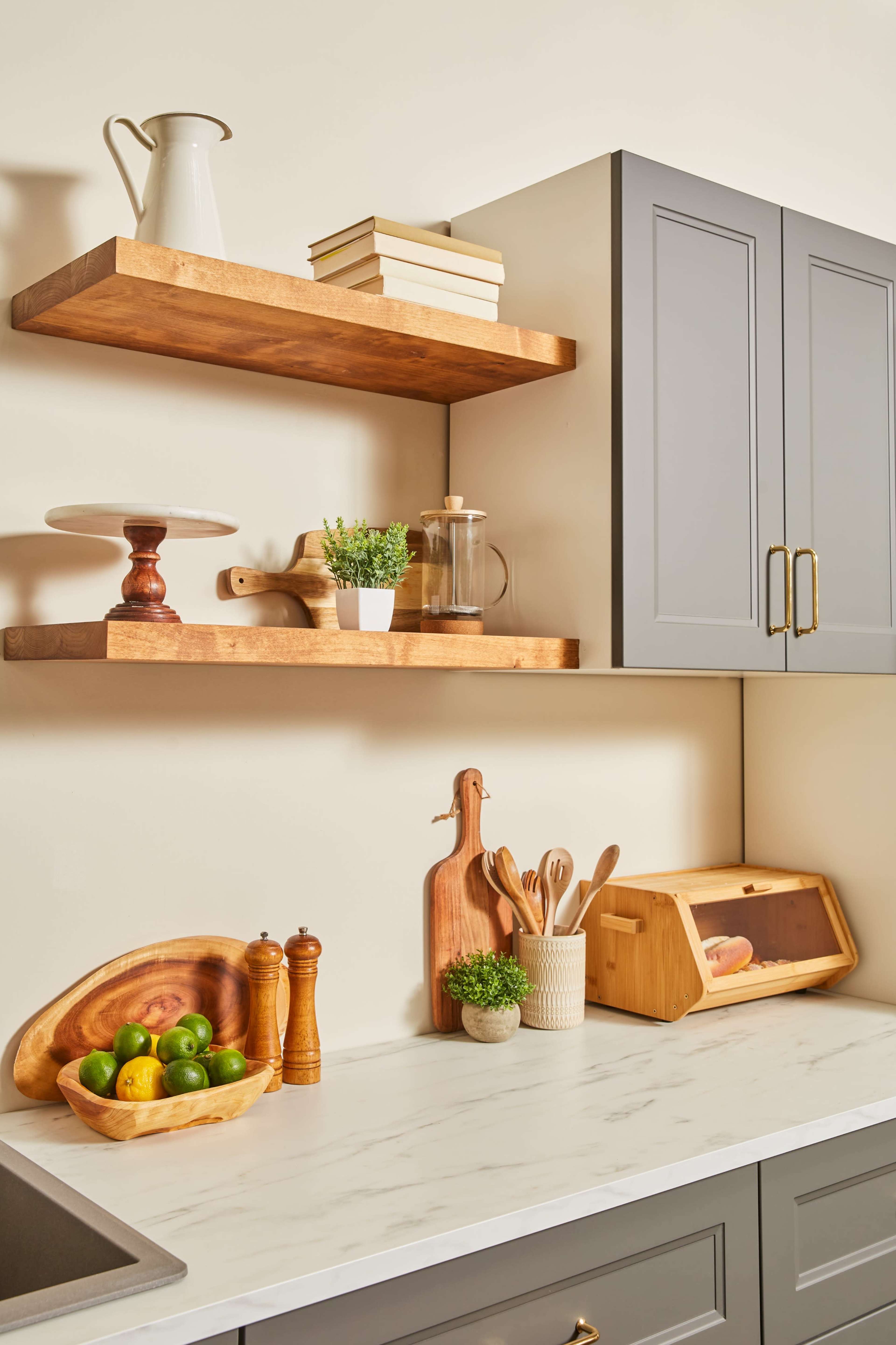 A modern kitchen displays wooden shelves holding cookbooks and decorative items above a marble countertop with various kitchen utensils and fresh produce.