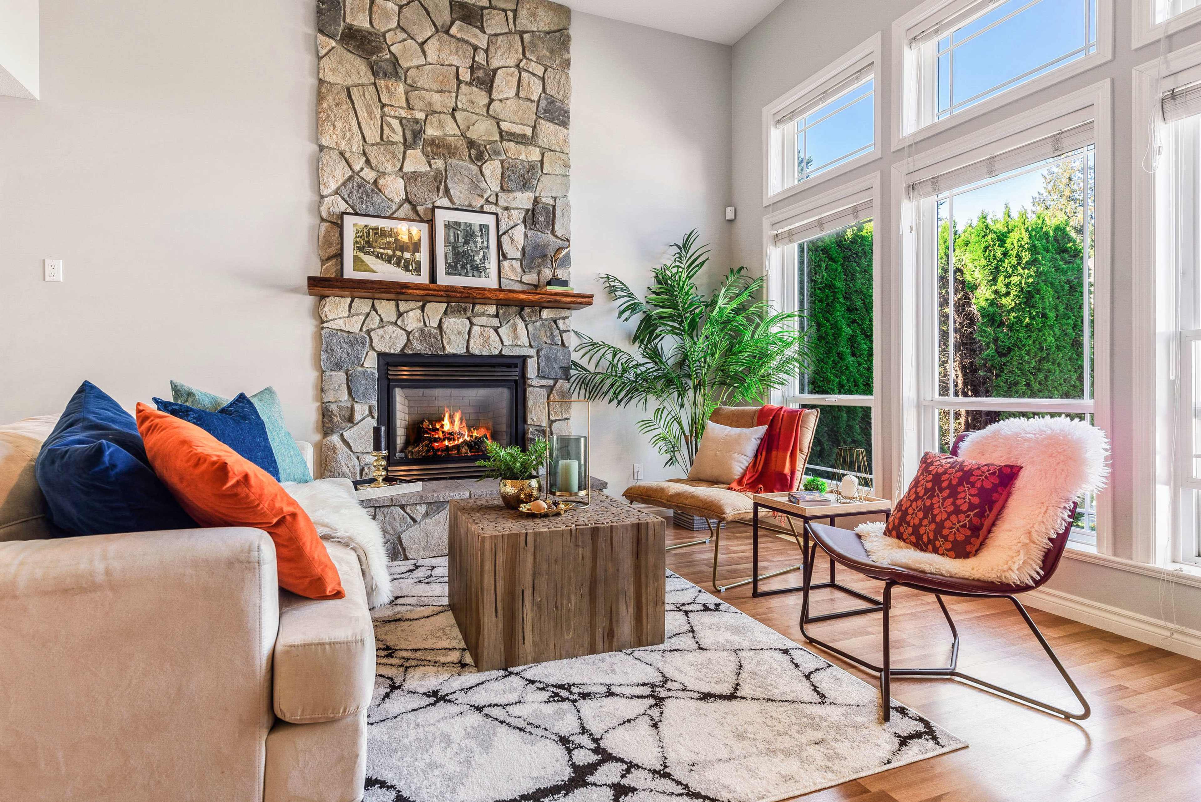 A cozy living room featuring a stone fireplace, a wooden coffee table, and a mix of colorful cushions on a sofa and chairs, with large windows providing natural light.
