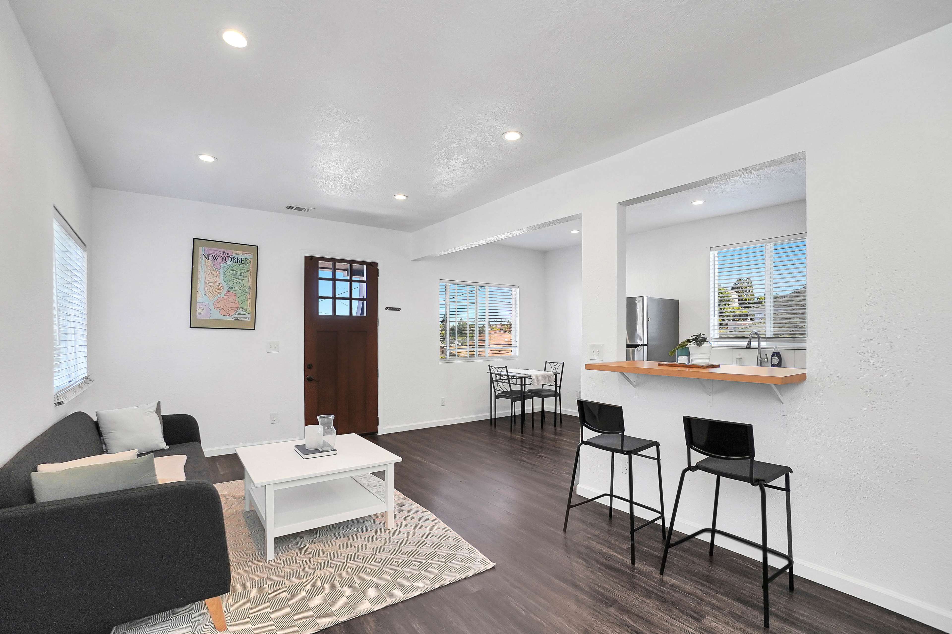 The image shows a modern, open-concept living space with a seating area, a small dining area, and a kitchen bar.