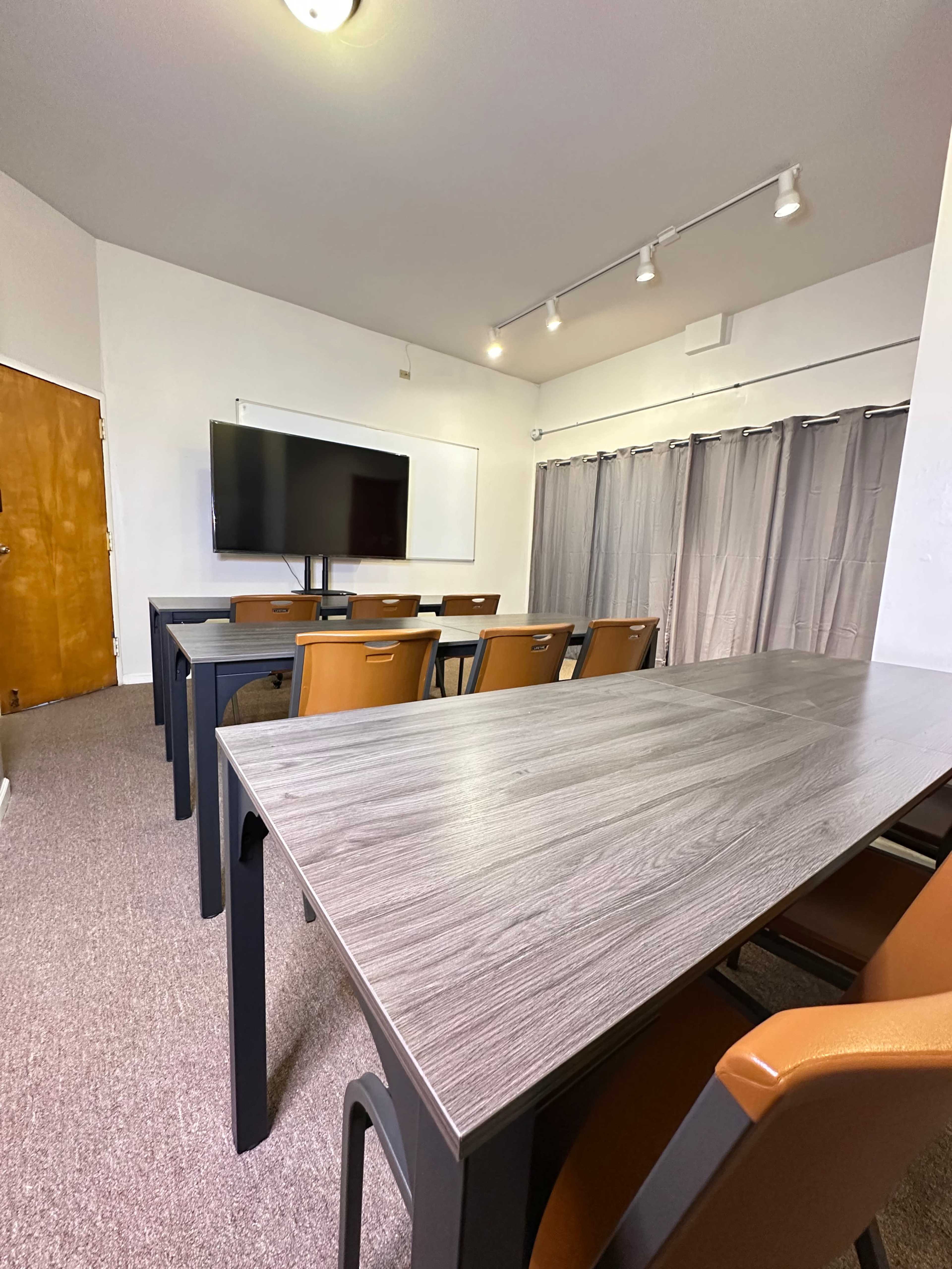 The image shows a conference room with a long table, several brown chairs, a television screen mounted on the wall, and gray curtains.