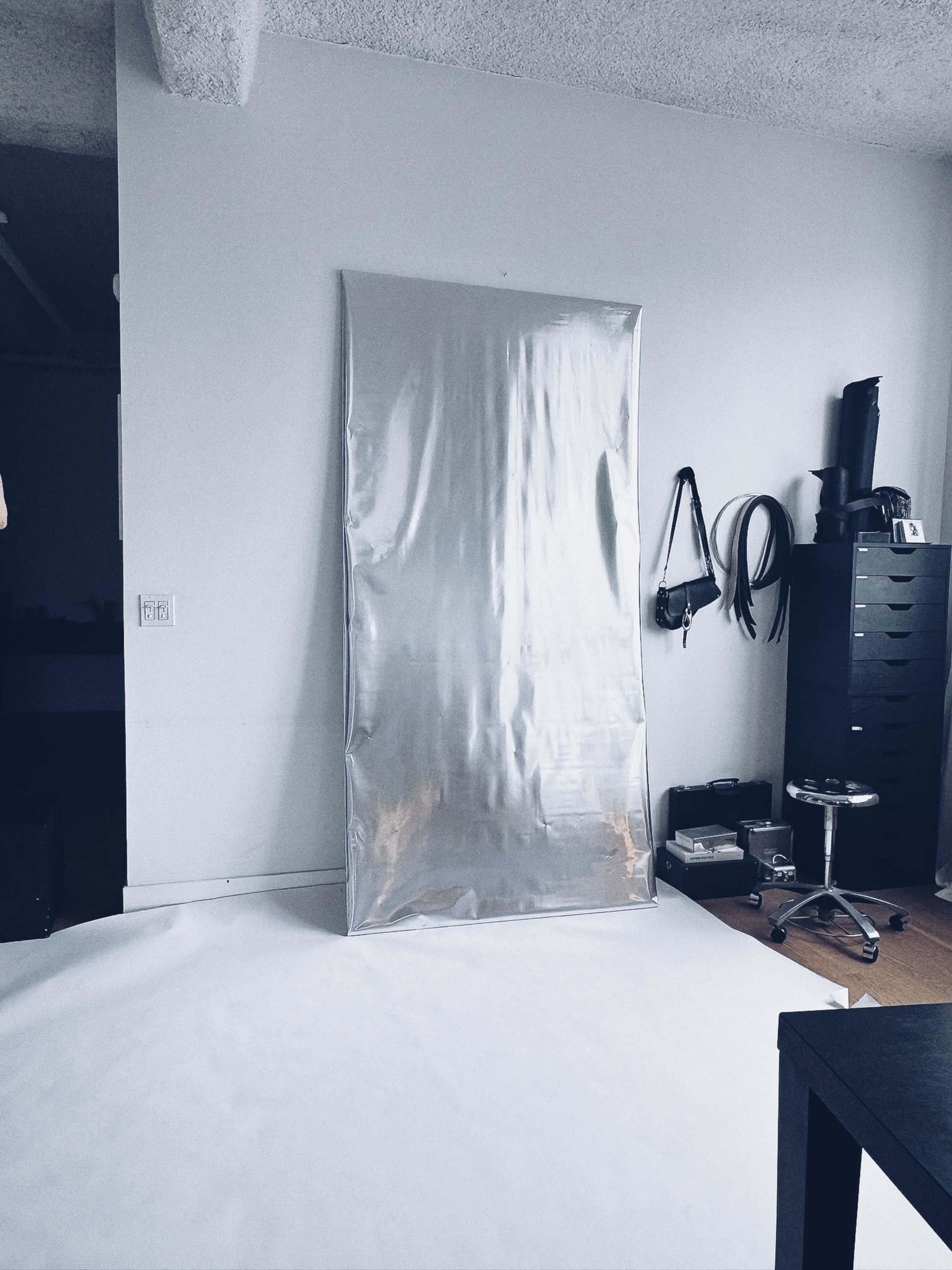The image shows a large, reflective silver panel mounted on a wall in a room with minimal furniture and a light-colored floor.
