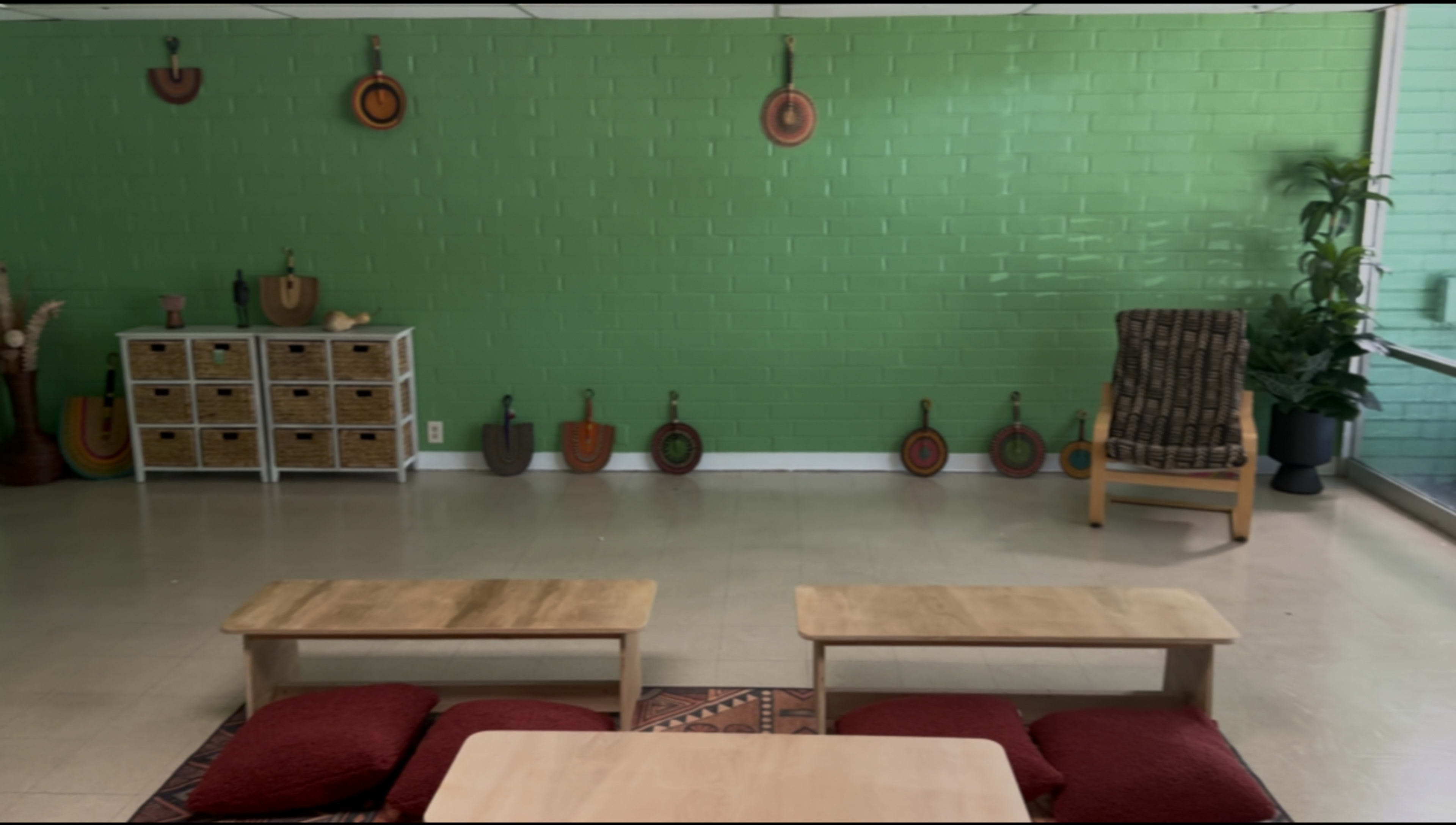 The image shows a room with green walls, two wooden tables, red floor cushions, and decorative items on the walls, including various shapes in a line along the floor.