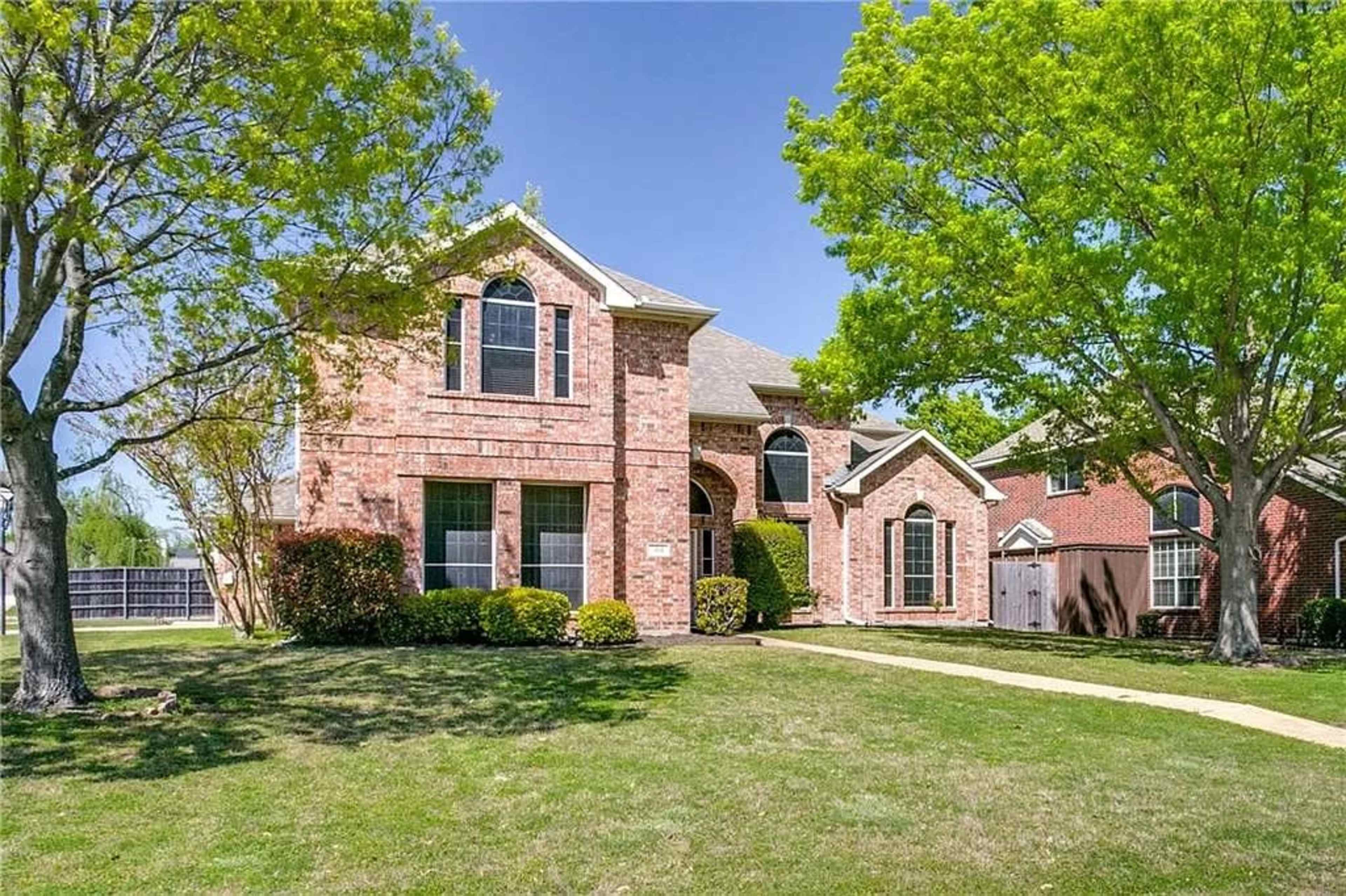 A two-story brick house with a landscaped front yard and large windows is situated on a green lawn.