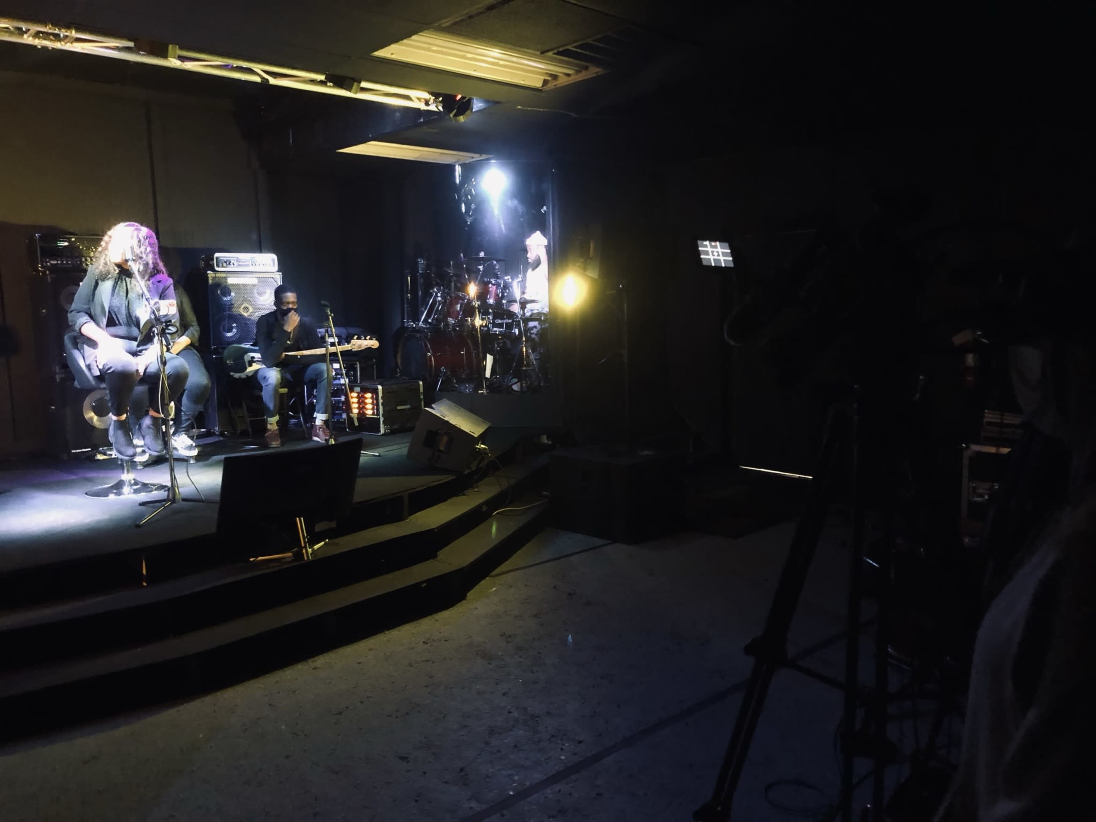 A musician plays guitar on stage while a drummer is visible in the background, and cameras are set up to capture the performance.