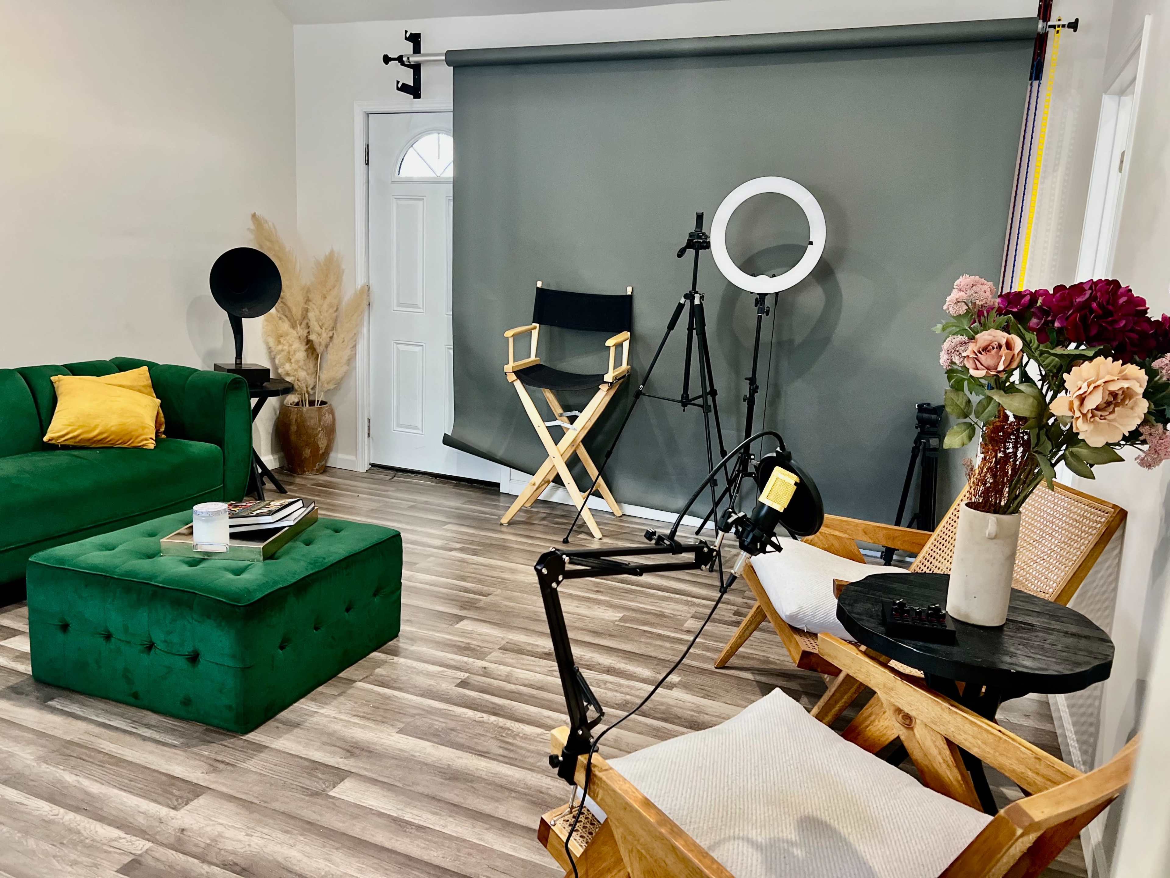A well-decorated room featuring a green velvet sofa, a matching ottoman, a director's chair, a ring light, and potted flowers, all arranged around a wooden table.
