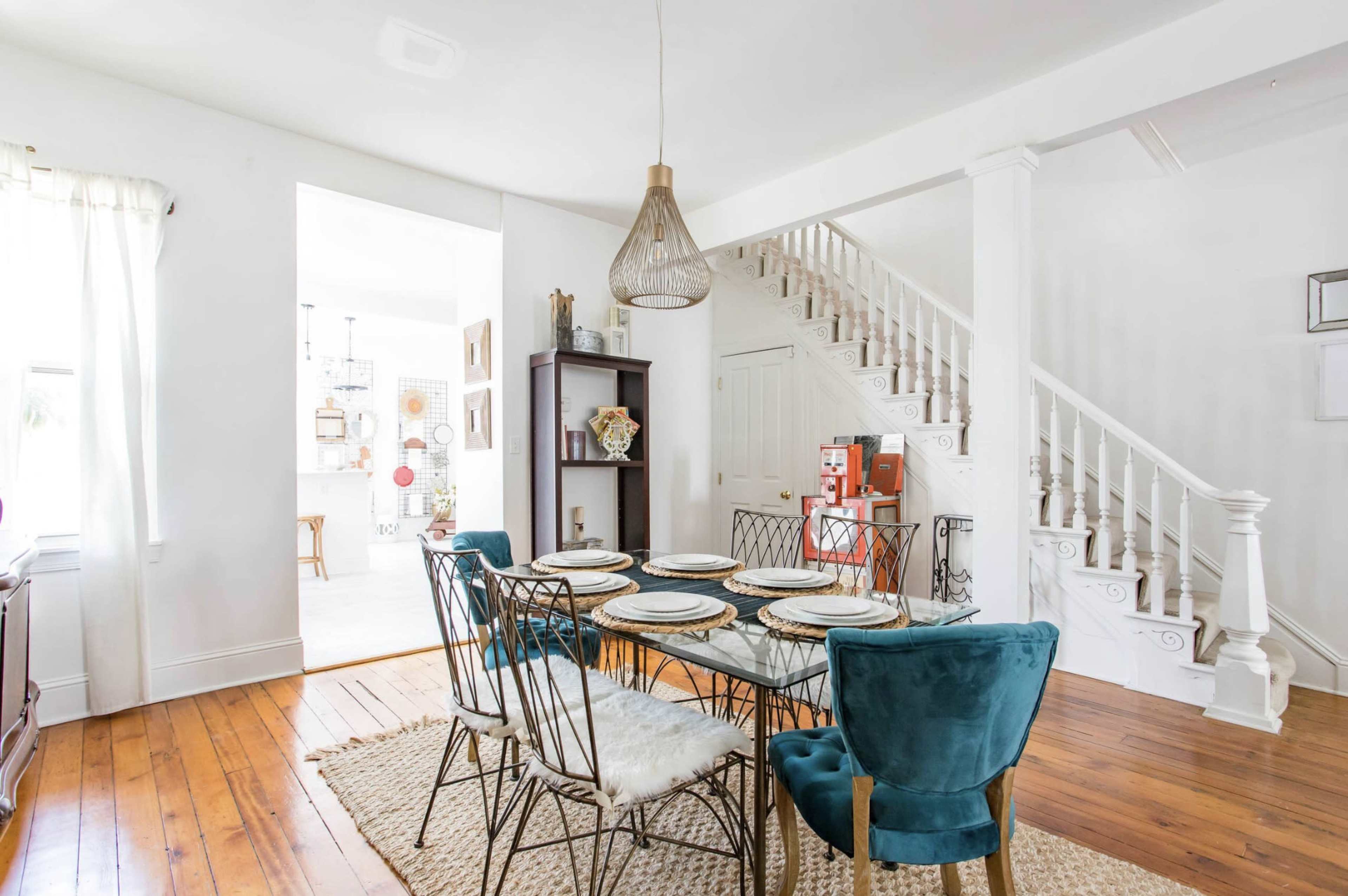 A dining area features a glass table with metal wire chairs, a woven rug, and a staircase in the background, leading to a well-lit space.