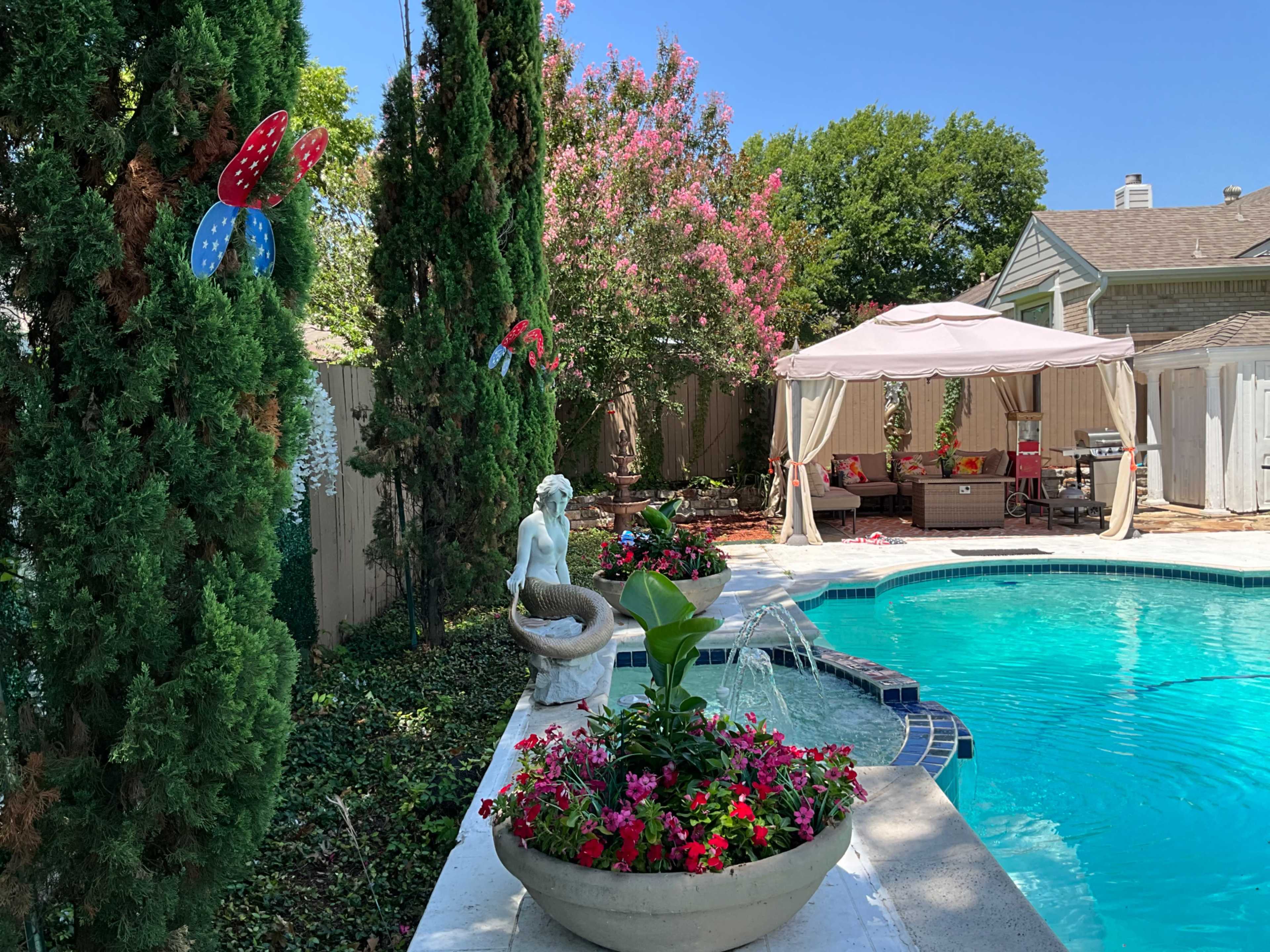 The image shows a swimming pool surrounded by colorful flower beds, a statue, and a shaded seating area with a canopy.