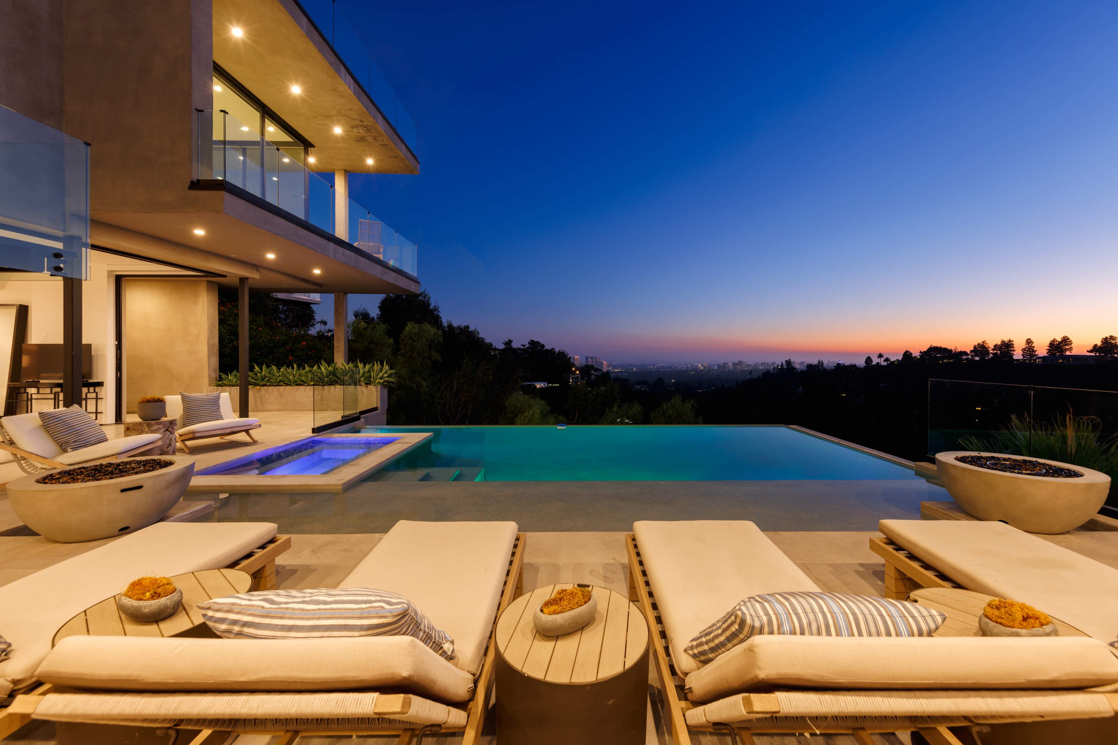 A modern villa with a pool and lounge chairs overlooking a scenic view at dusk.