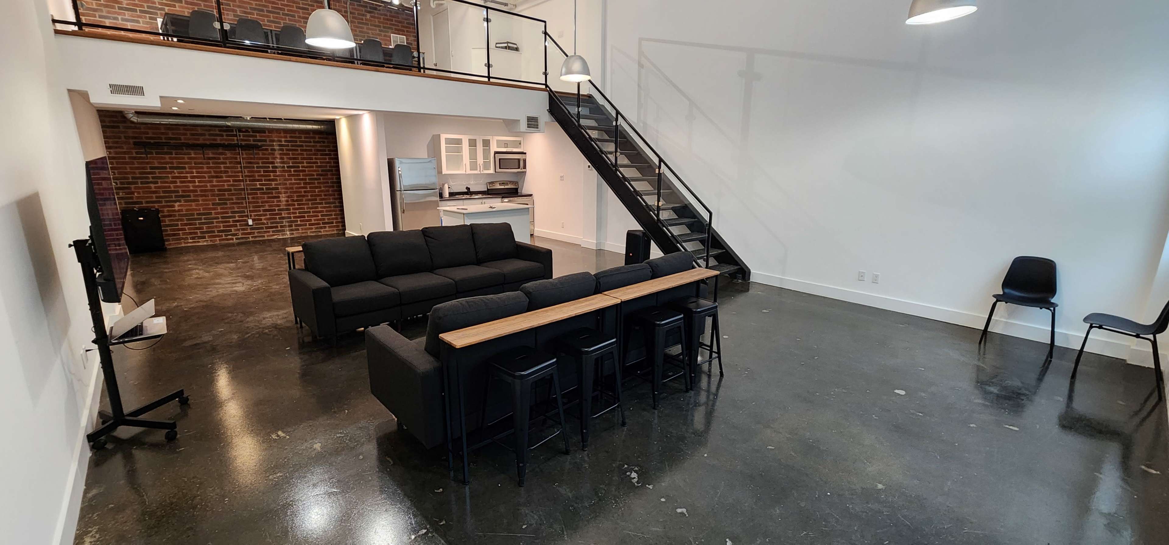 A spacious, modern loft features a black sectional sofa with a wooden tabletop, a metal staircase, and a kitchen area visible in the background.