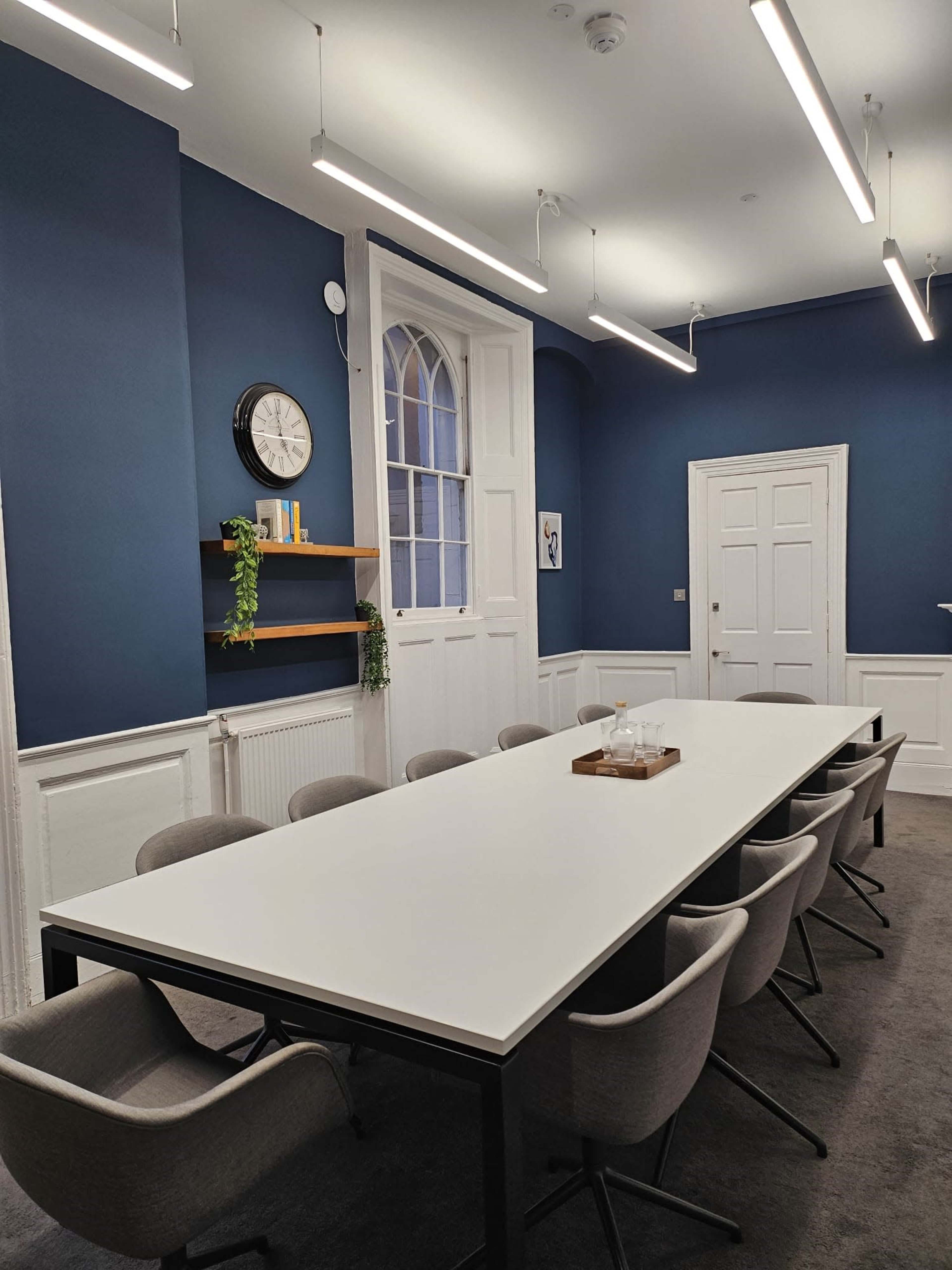 A long white table is surrounded by gray chairs in a blue-walled conference room featuring a clock and shelves with decorative items.
