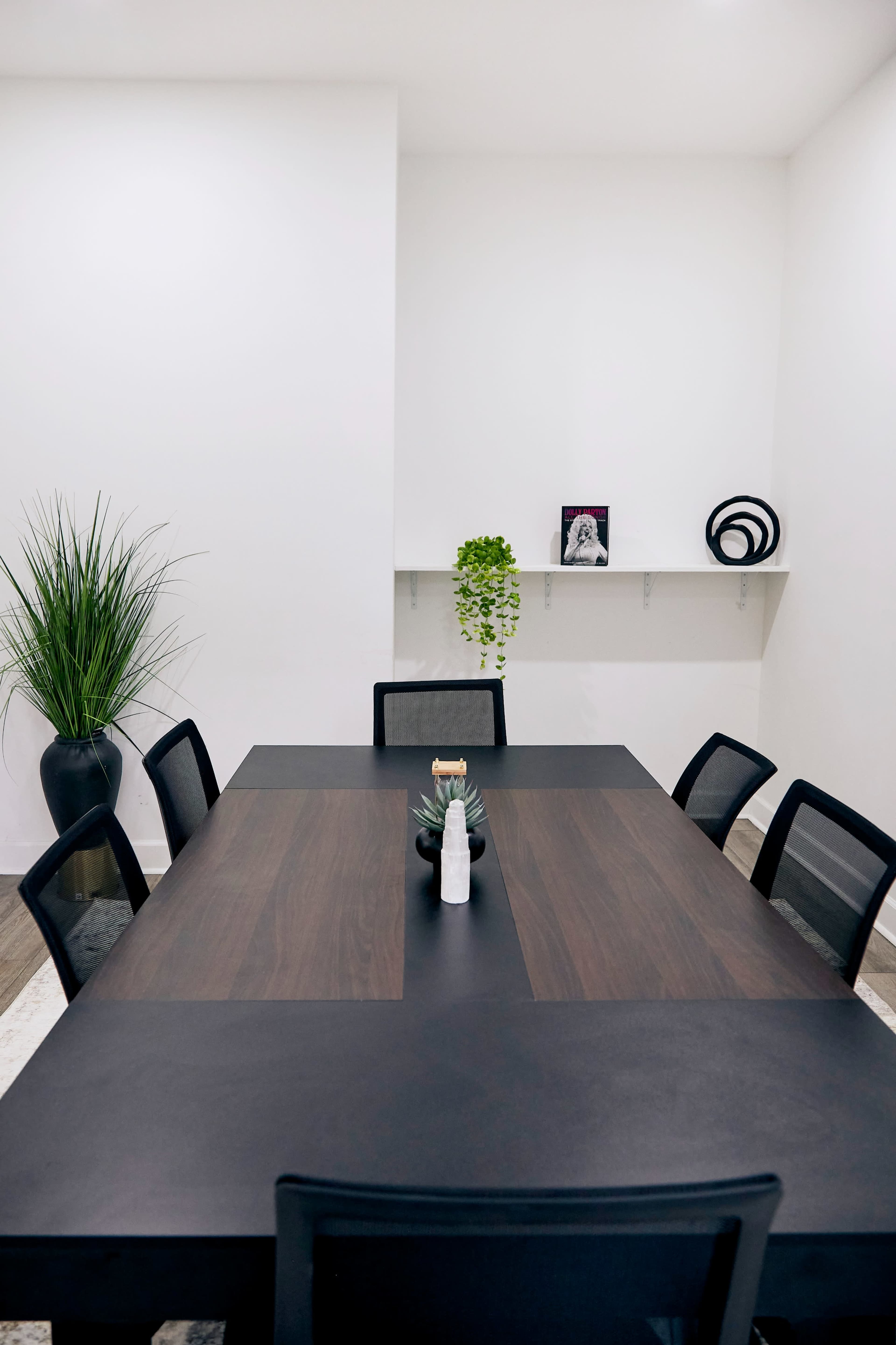 A modern conference room features a large black table surrounded by black chairs, with a potted plant and decorative items on the shelves and walls.