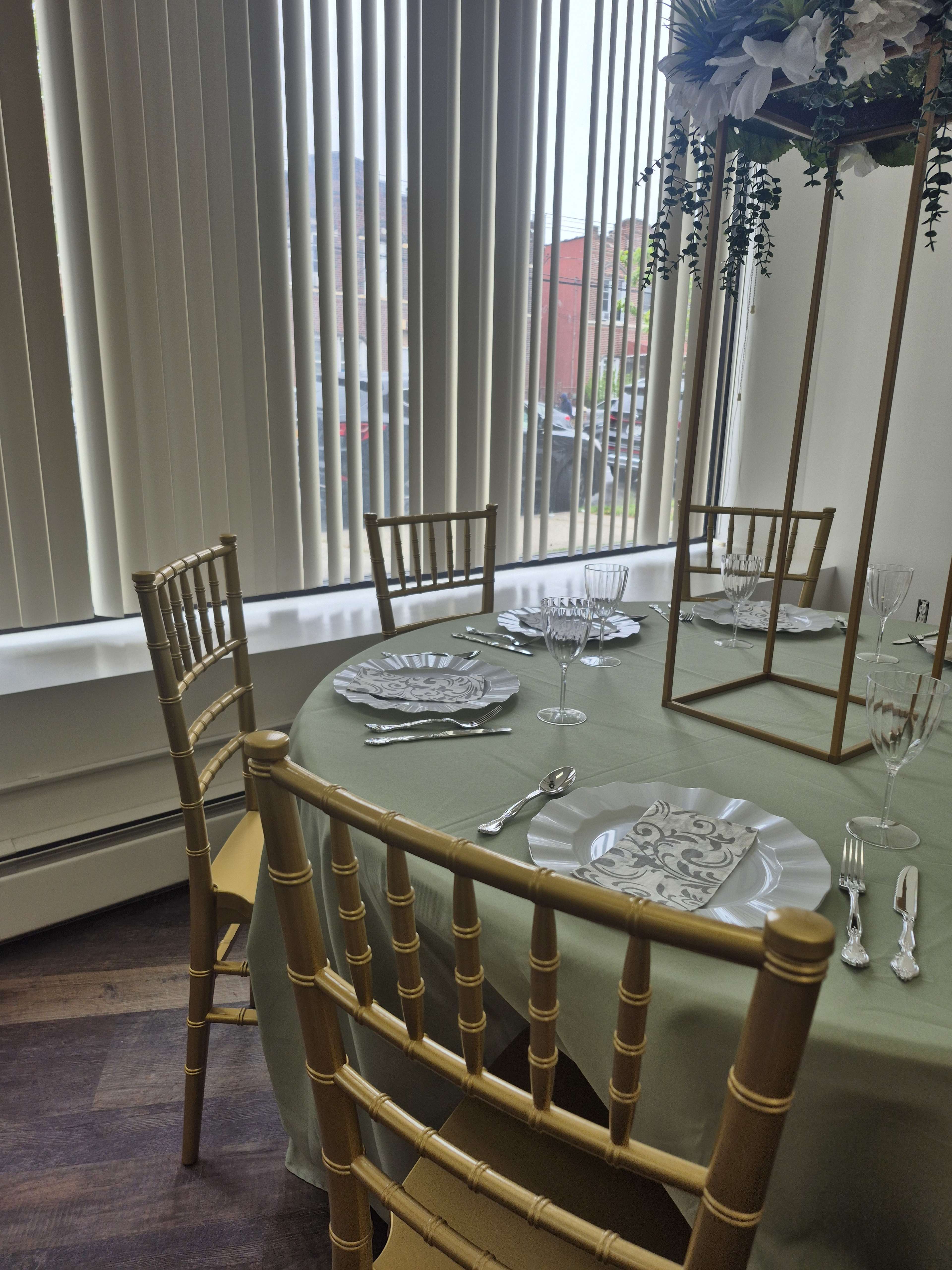 A round table is set with silver utensils and patterned plates, surrounded by gold chiavari chairs in a brightly lit room with large windows.