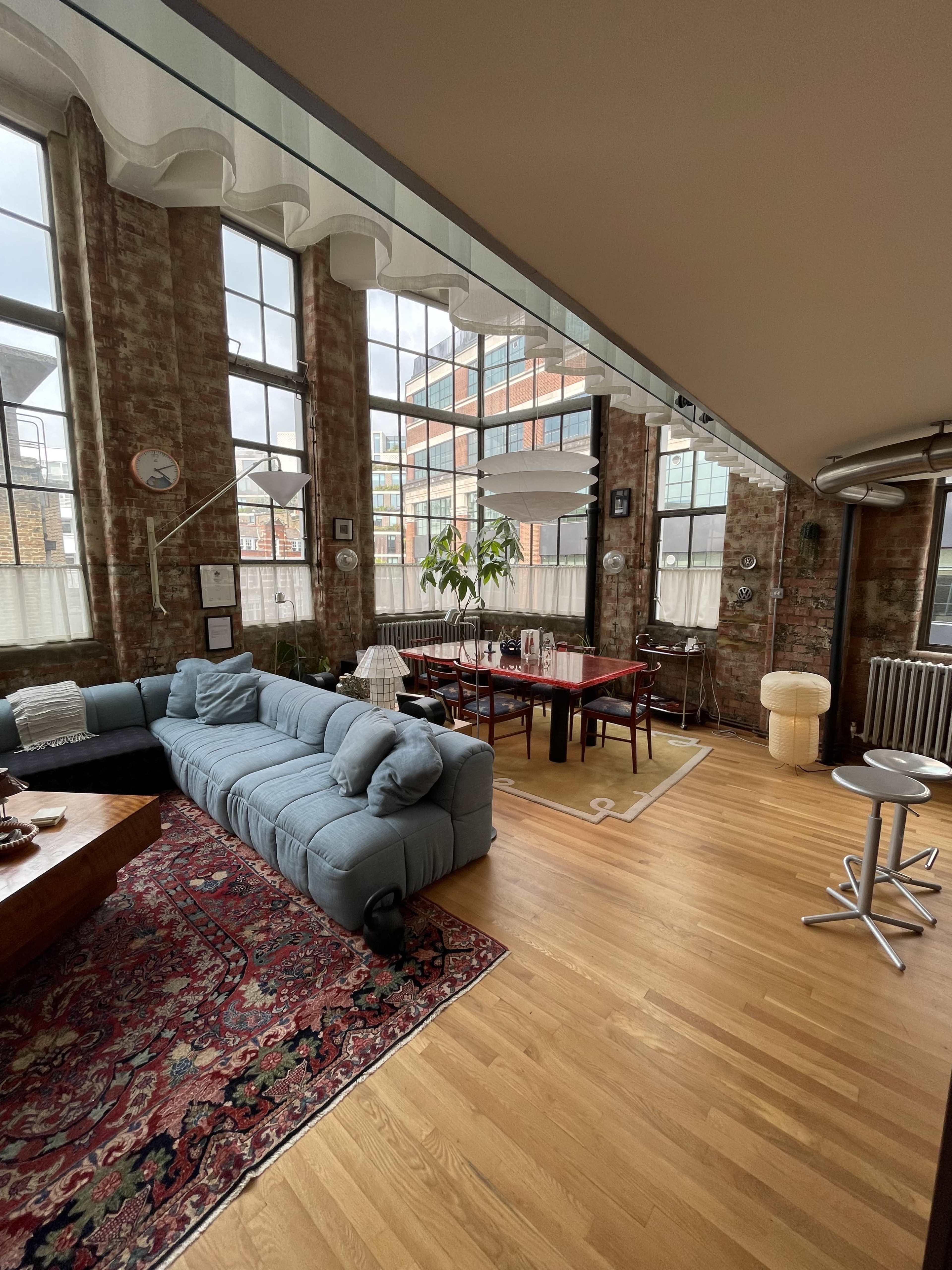 The image shows a spacious living area with exposed brick walls, a large light blue sectional sofa, a dining table set for several people, and plenty of natural light from large windows.