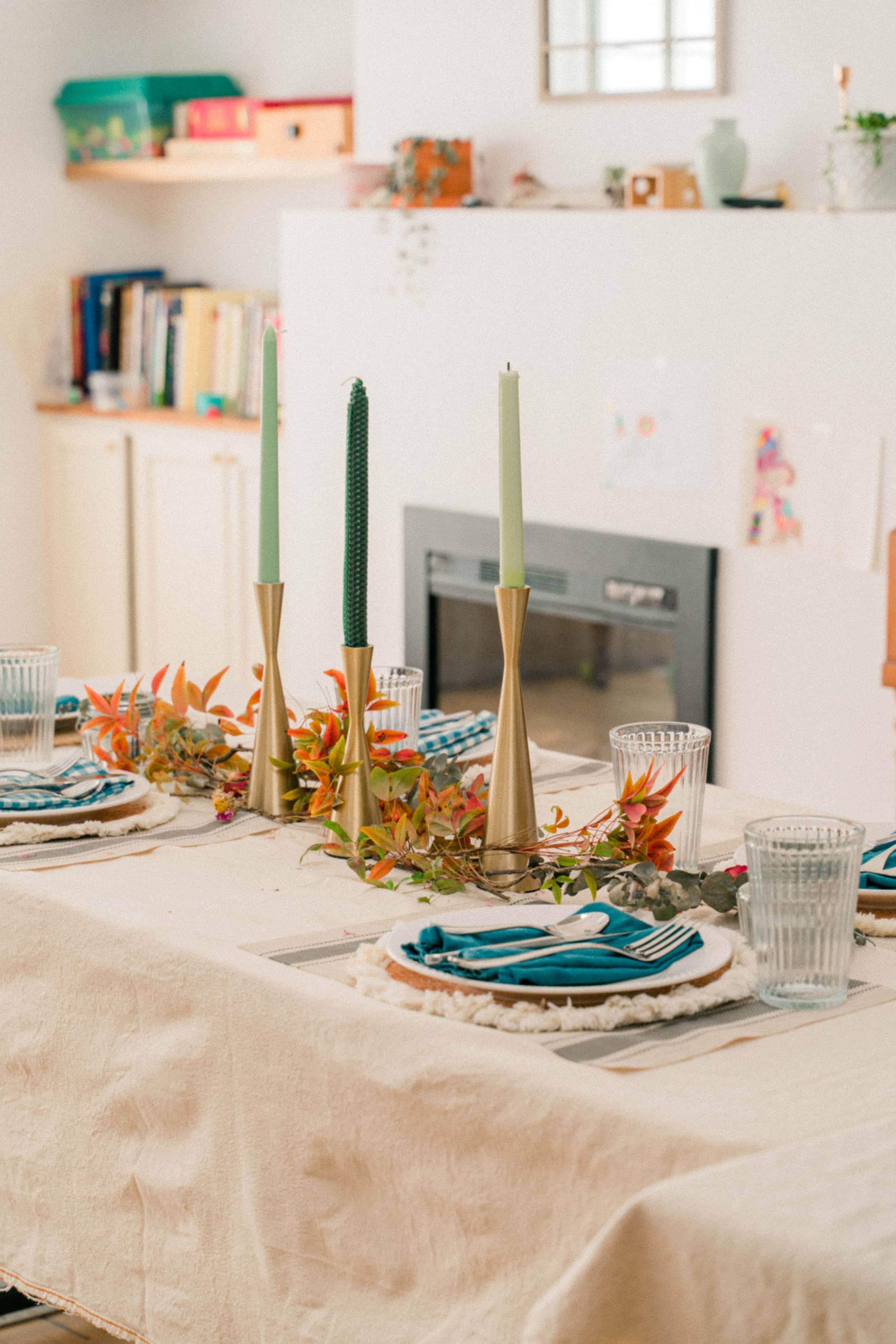 A dining table is set with decorative plates, cutlery, and candles, surrounded by a neatly arranged room with shelves and decor in the background.