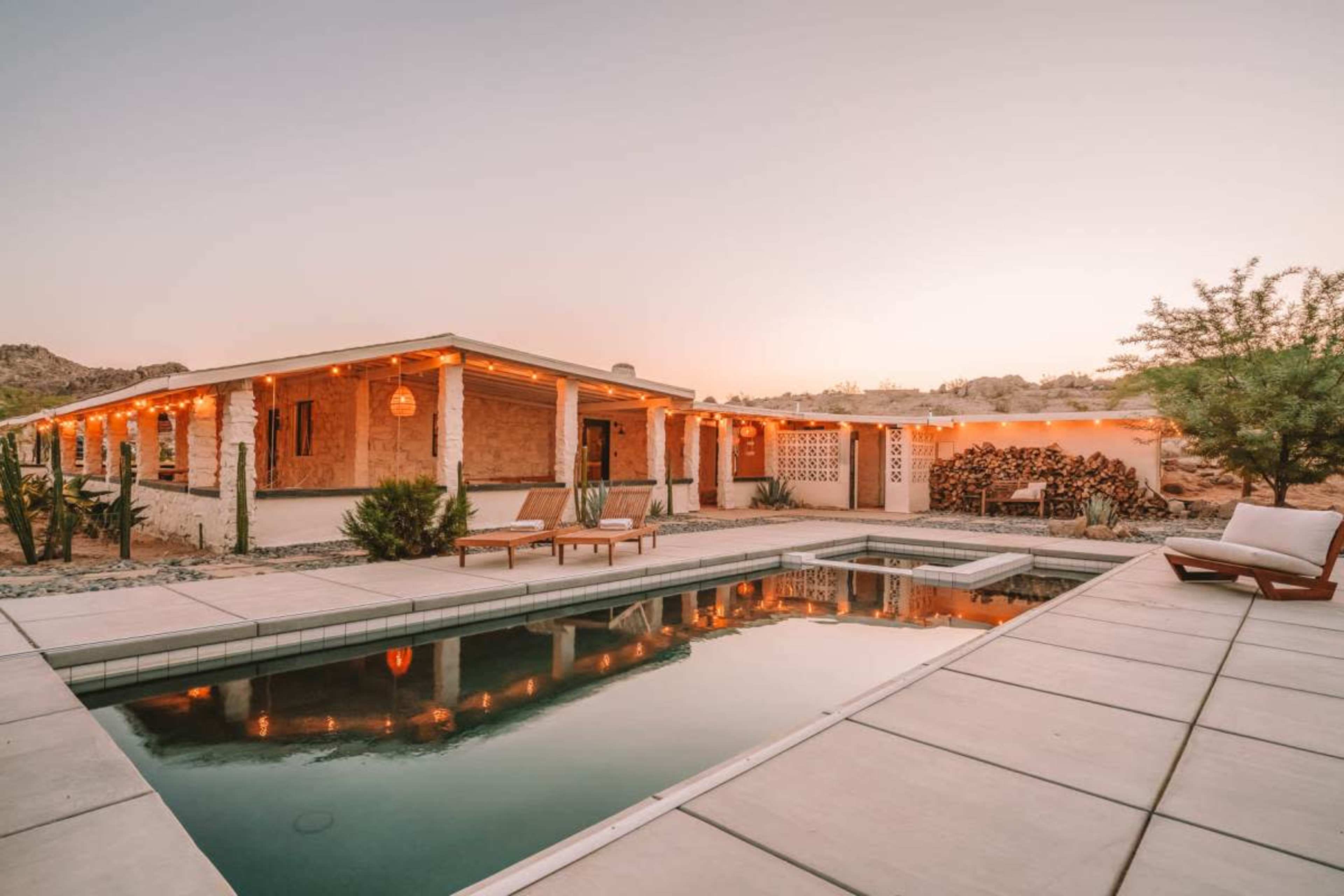 A desert house with a swimming pool is illuminated by string lights as the sun sets in the background.