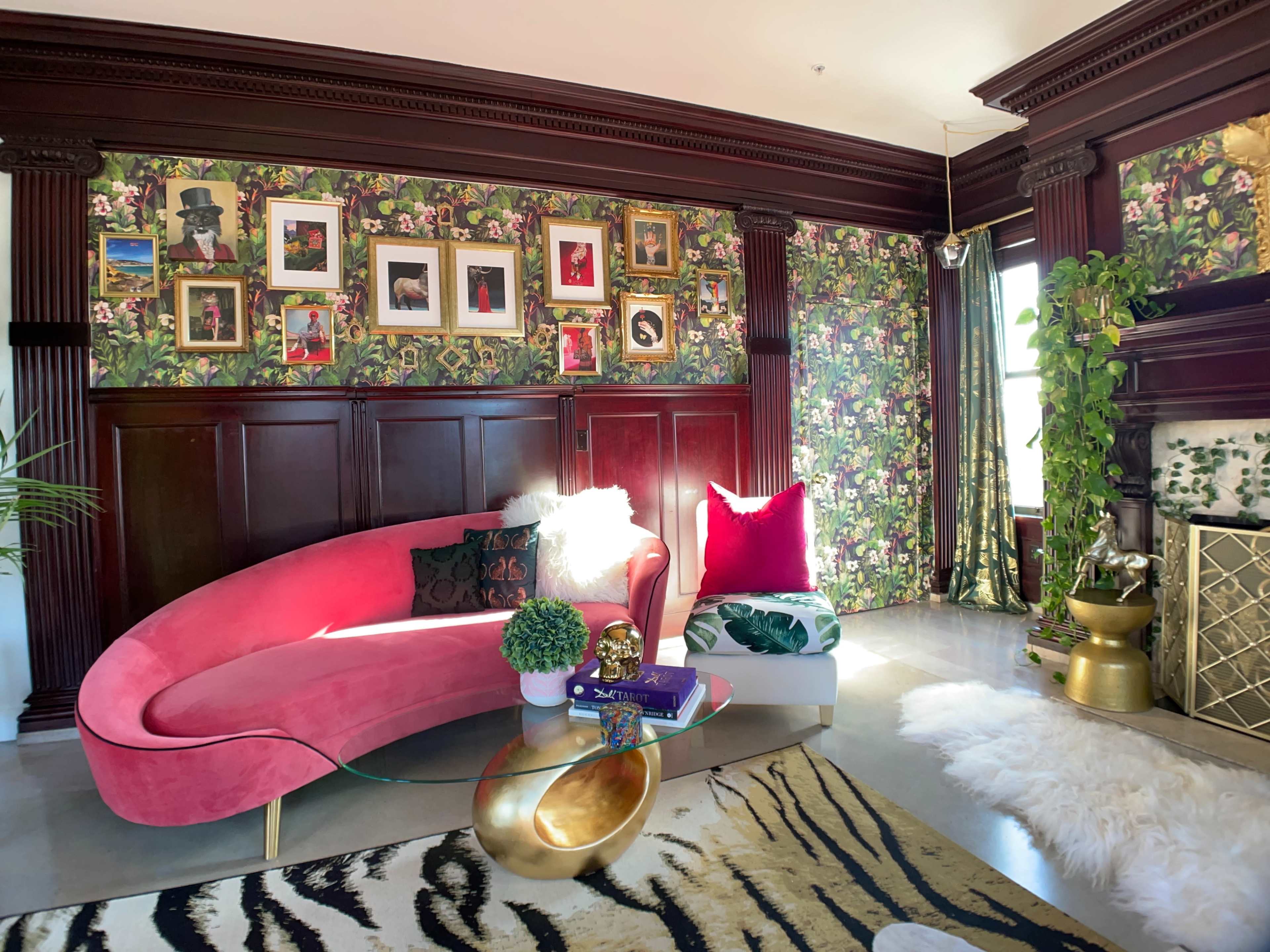 The interior features a pink curved sofa, a glass coffee table, and decorative wallpaper adorned with floral patterns and framed artwork.
