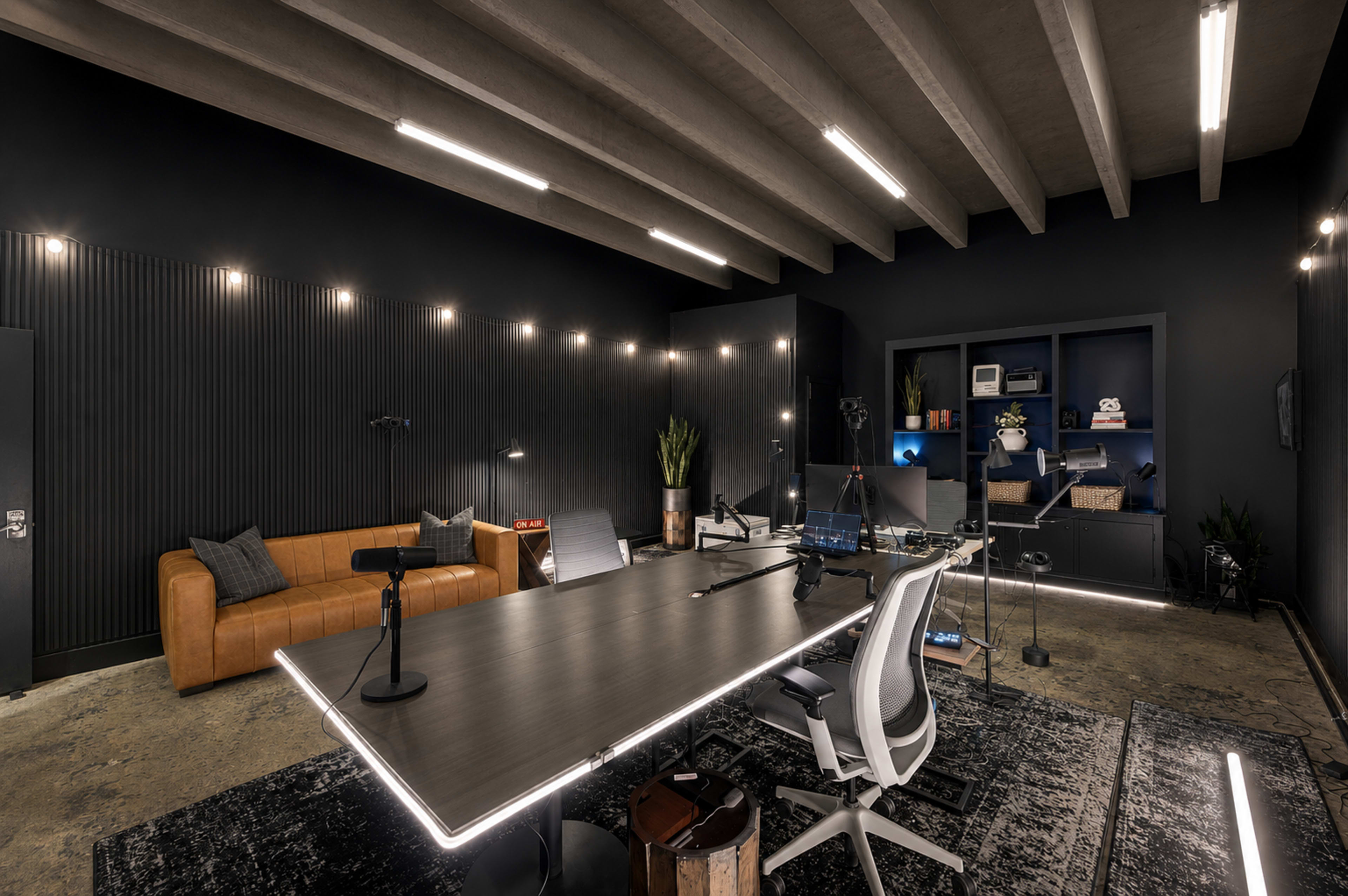 The image shows a modern office space with a large table, microphones, a leather couch, and shelves against dark walls, illuminated by strip lighting.