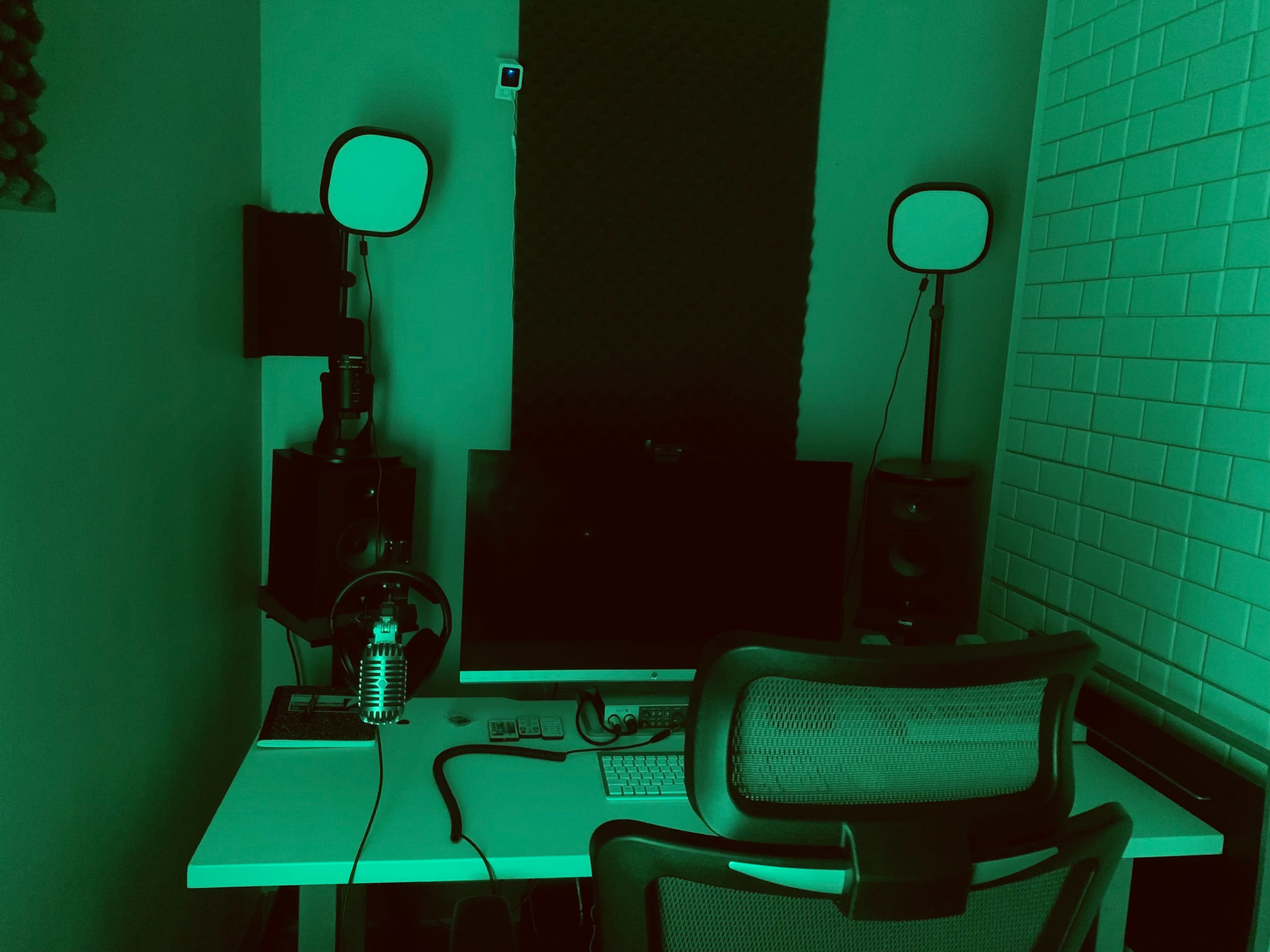 A small, dimly lit workspace featuring a computer monitor, speakers, and a chair, all surrounded by a green light.