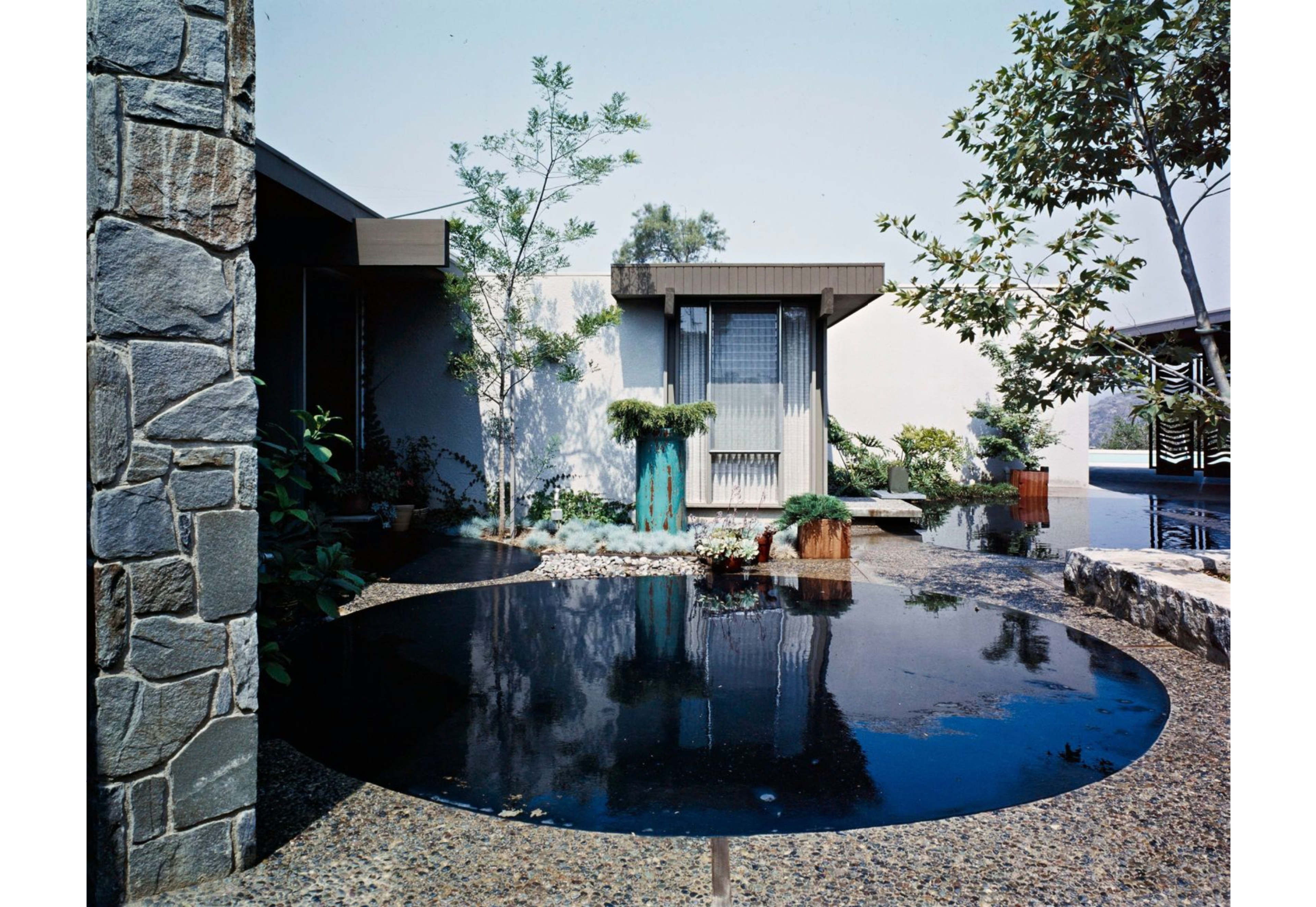 The image depicts a modern architectural setting featuring a serene pond surrounded by greenery and stone accents.