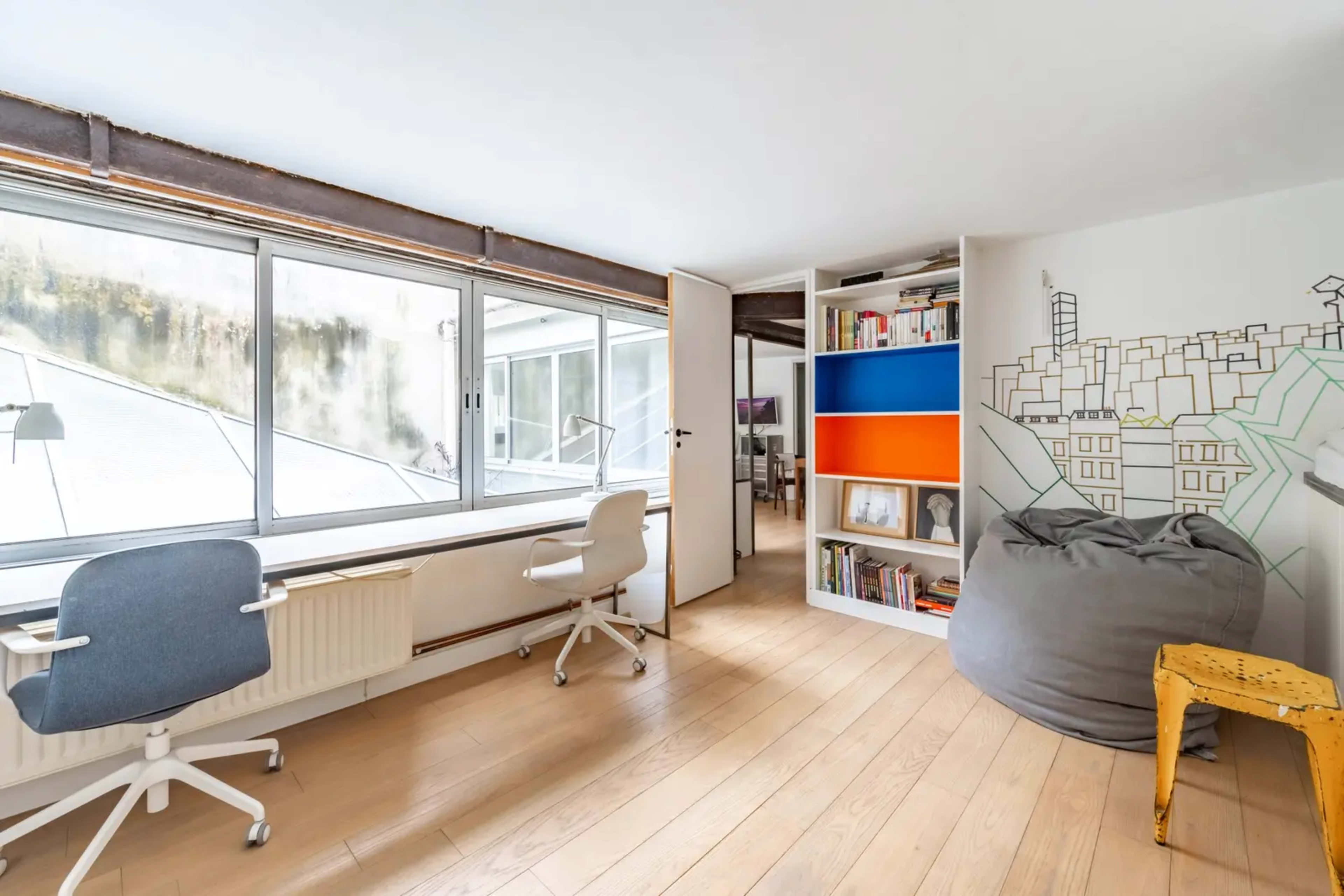 A bright, minimalist workspace features a large window, wooden floors, and modern furniture, with a colorful bookshelf and a bean bag in one corner.