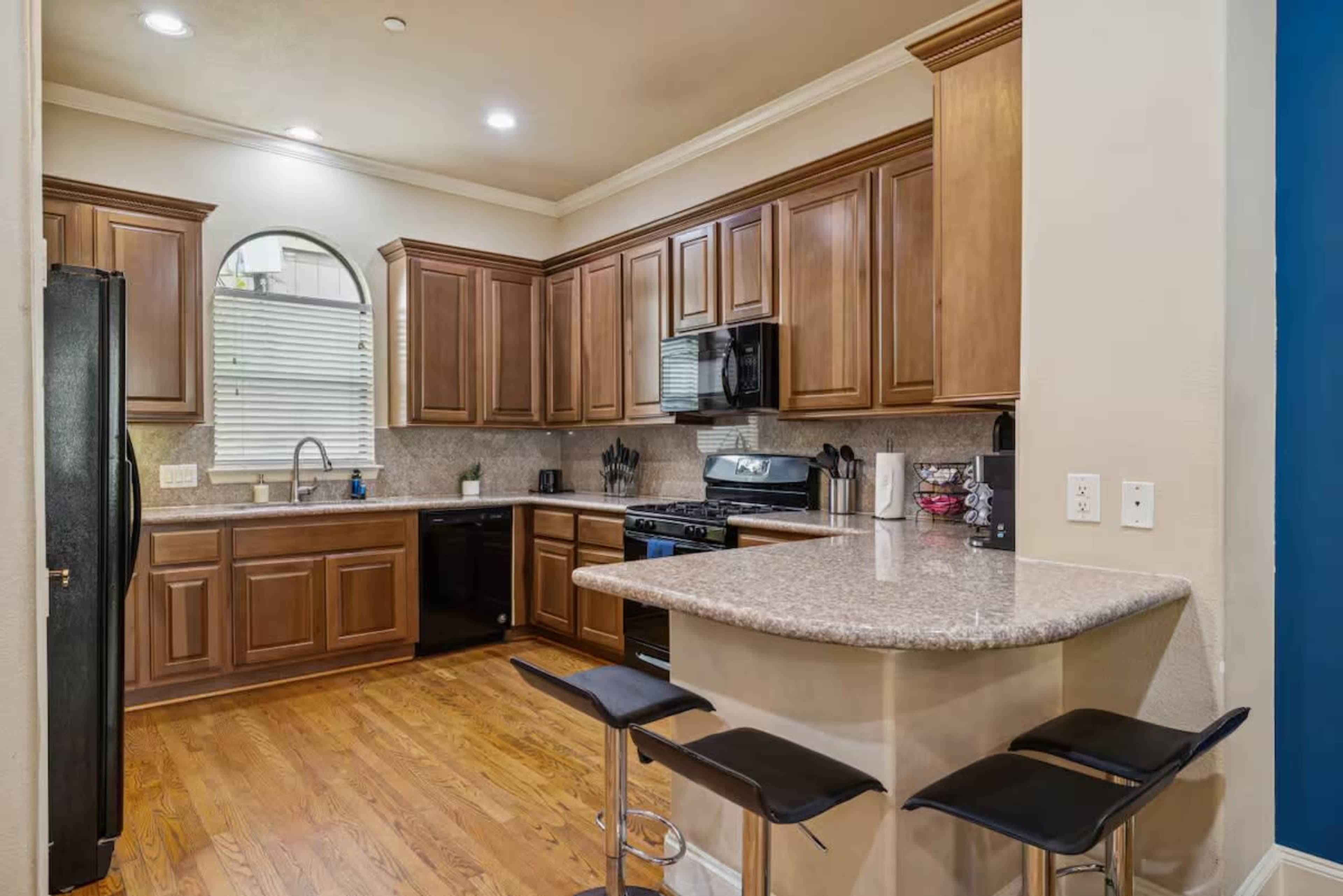 The image shows a modern kitchen featuring wooden cabinets, a granite countertop with bar stools, and stainless steel appliances.