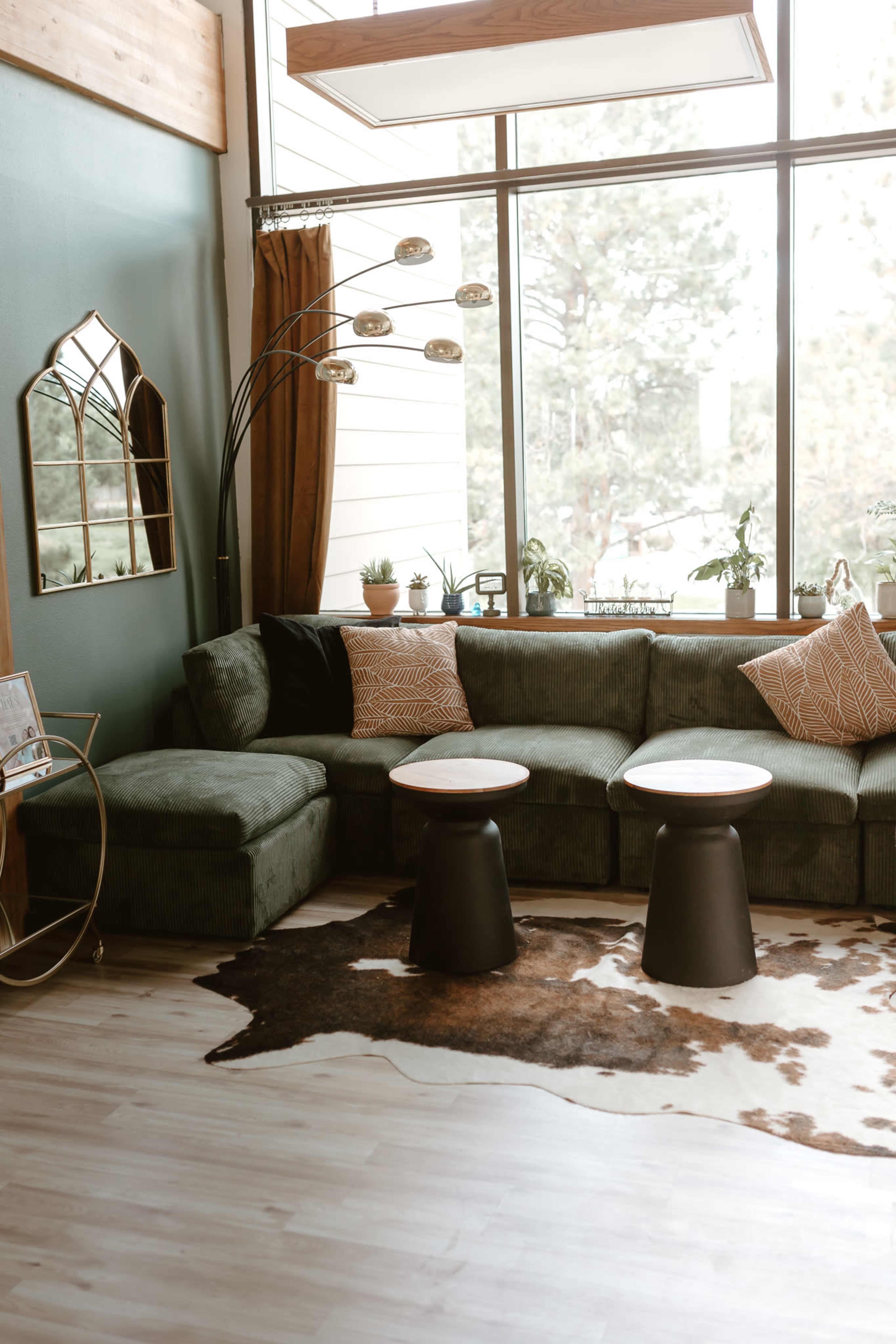 A cozy living room features a green sectional sofa, two round black tables, and large windows with indoor plants and natural light.