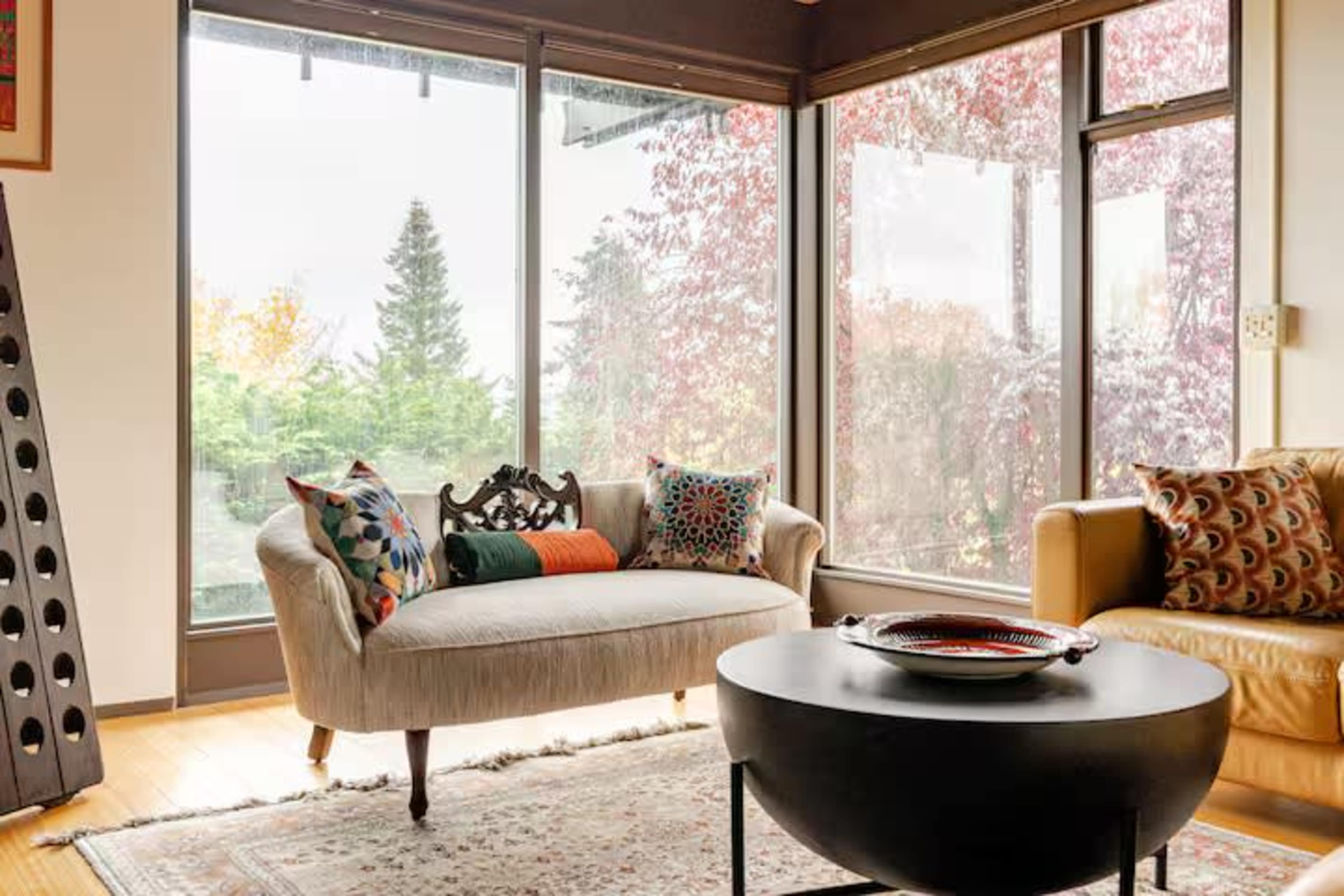 A cozy living room features a light-colored sofa with patterned cushions and a brown leather chair, surrounded by large windows that showcase a vibrant outdoor landscape.