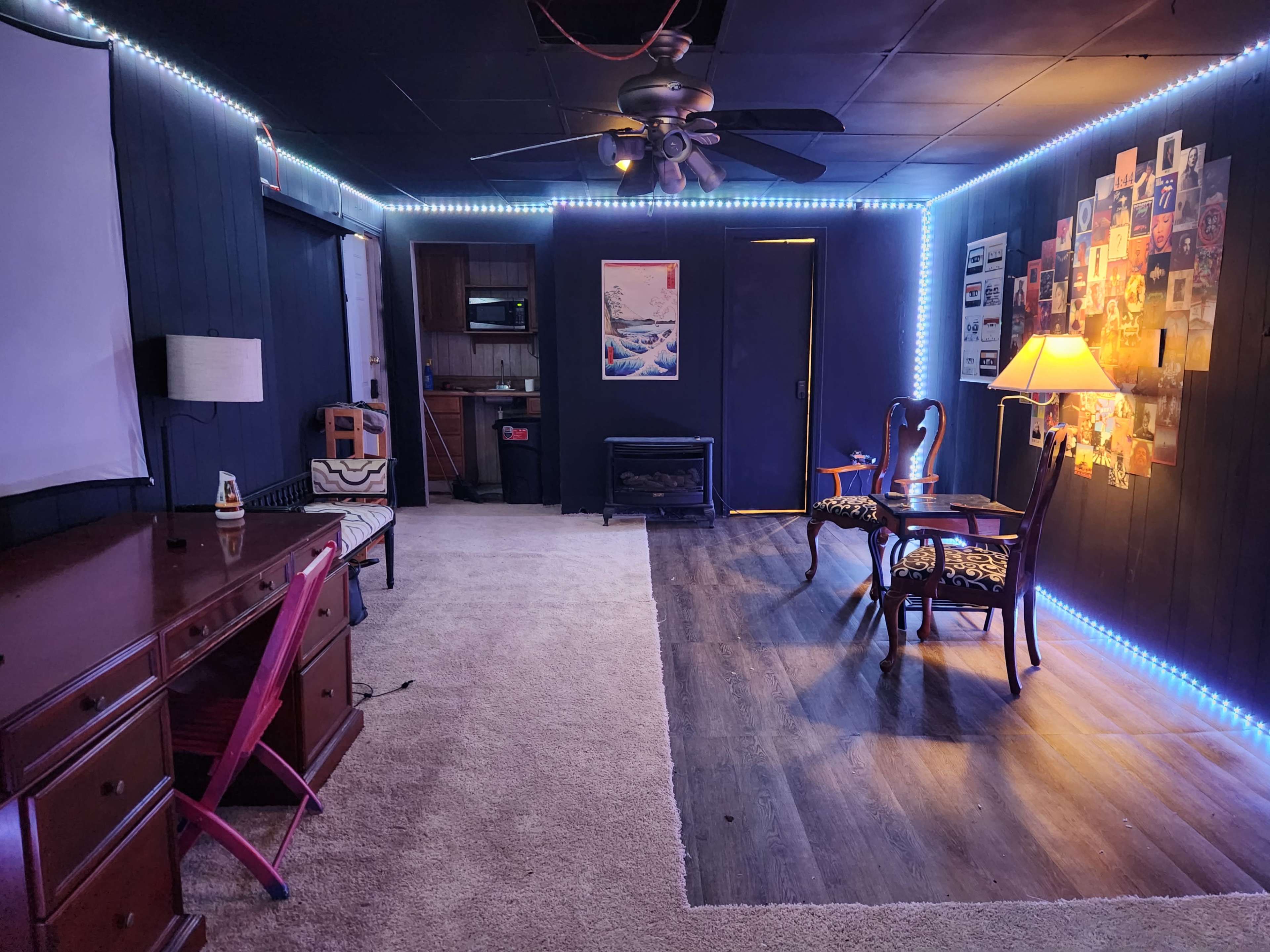 The image shows a room with a carpeted floor, featuring a desk with a pink chair, two armchairs, a lamp, a kitchenette area, and walls illuminated by LED strip lighting.