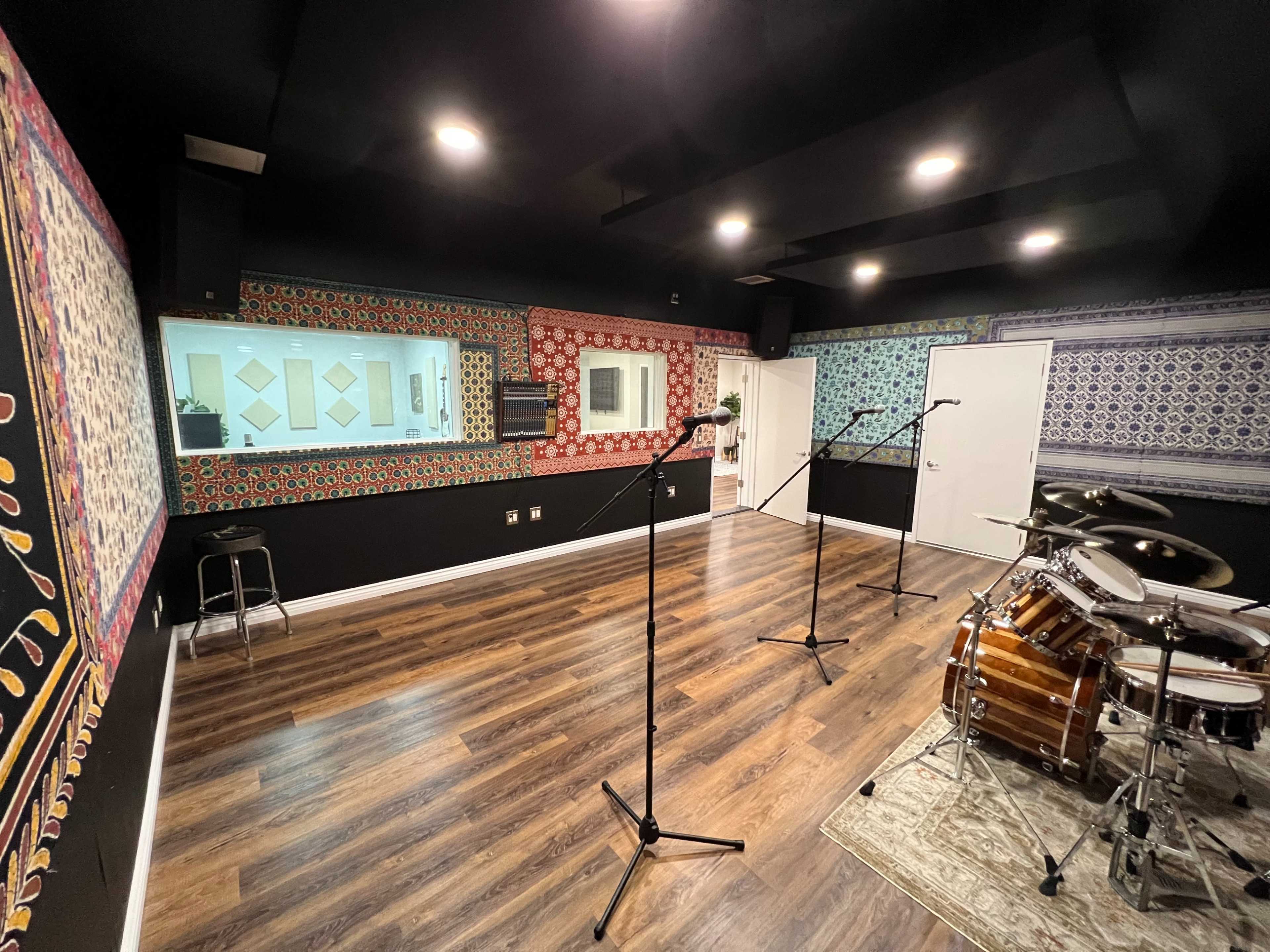 The image shows a recording studio with patterned walls, several microphone stands positioned for use, and a drum set on a rug in the corner.
