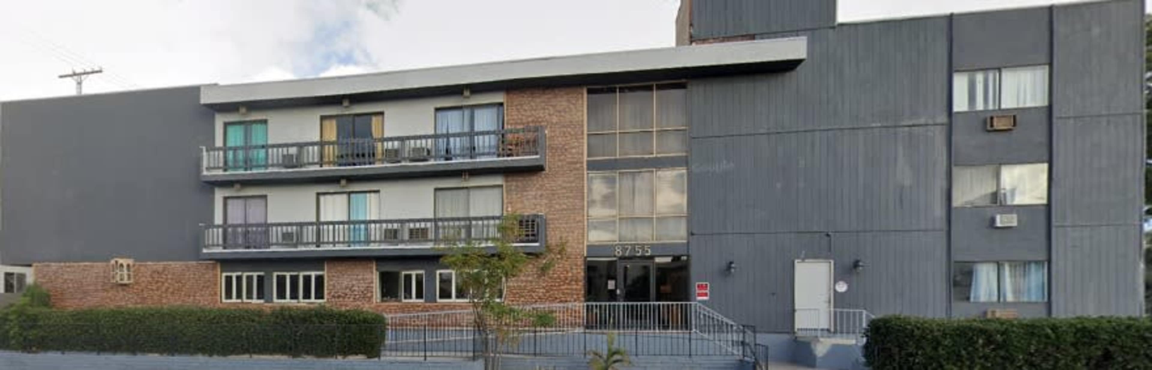 The image shows a modern multi-story building with a mix of brick and gray paneling, featuring balconies and large windows.