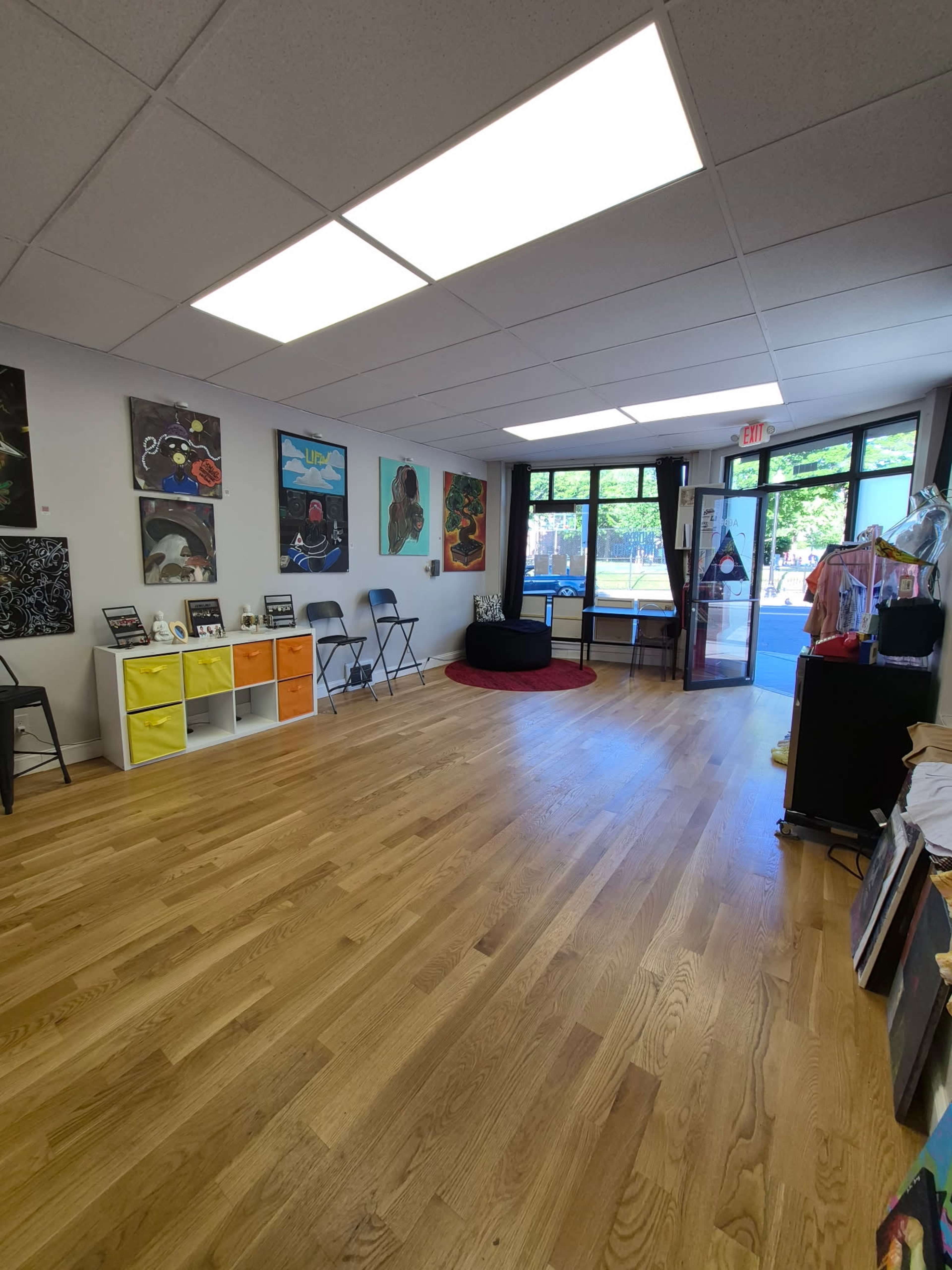 The image shows a spacious room with wooden flooring, featuring colorful storage units, chairs, artwork on the walls, and large windows that open to an outdoor area.