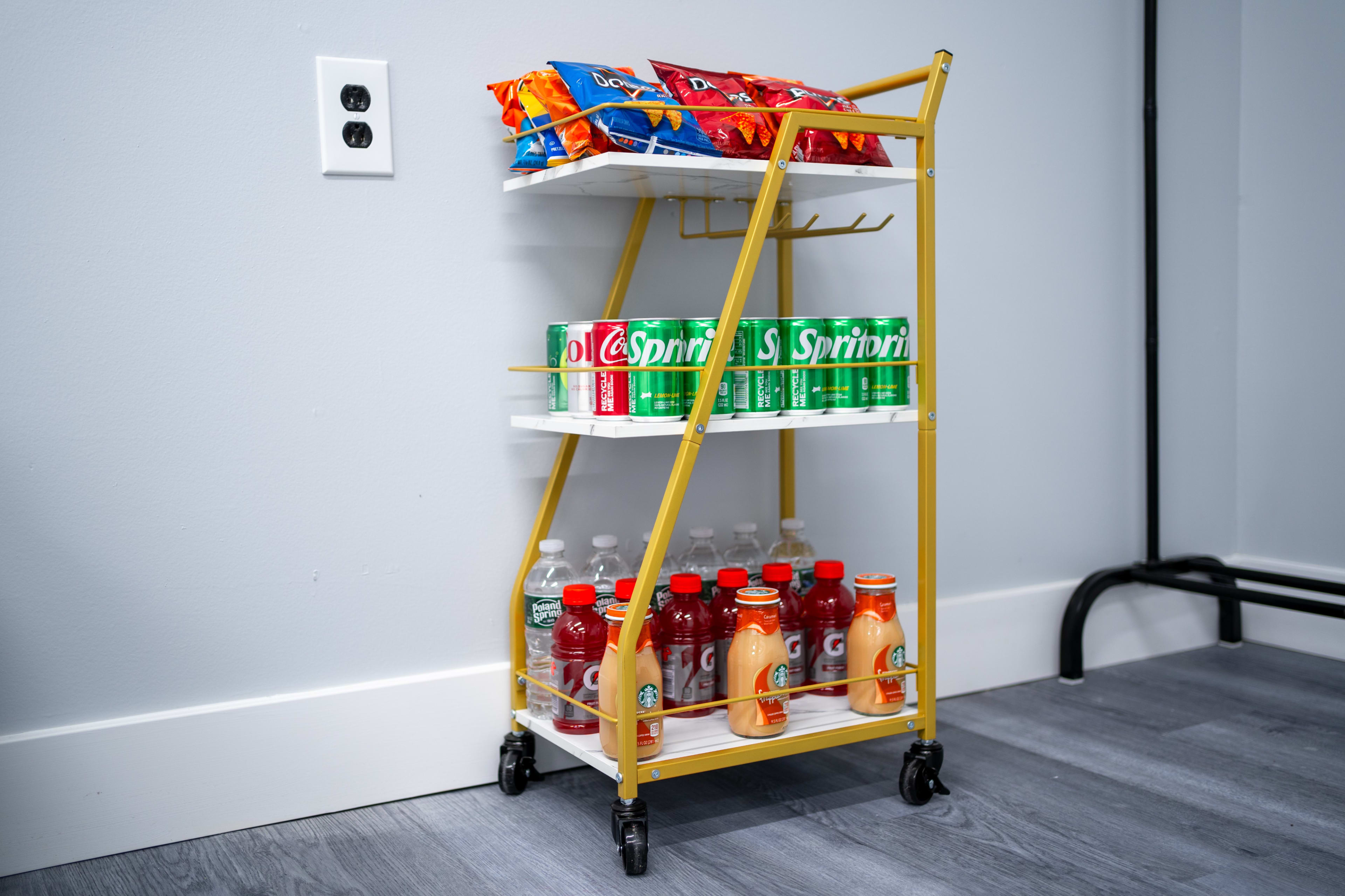 A gold metal snack cart with three shelves, stocked with snack bags, bottled drinks, and various beverages, positioned against a gray wall.